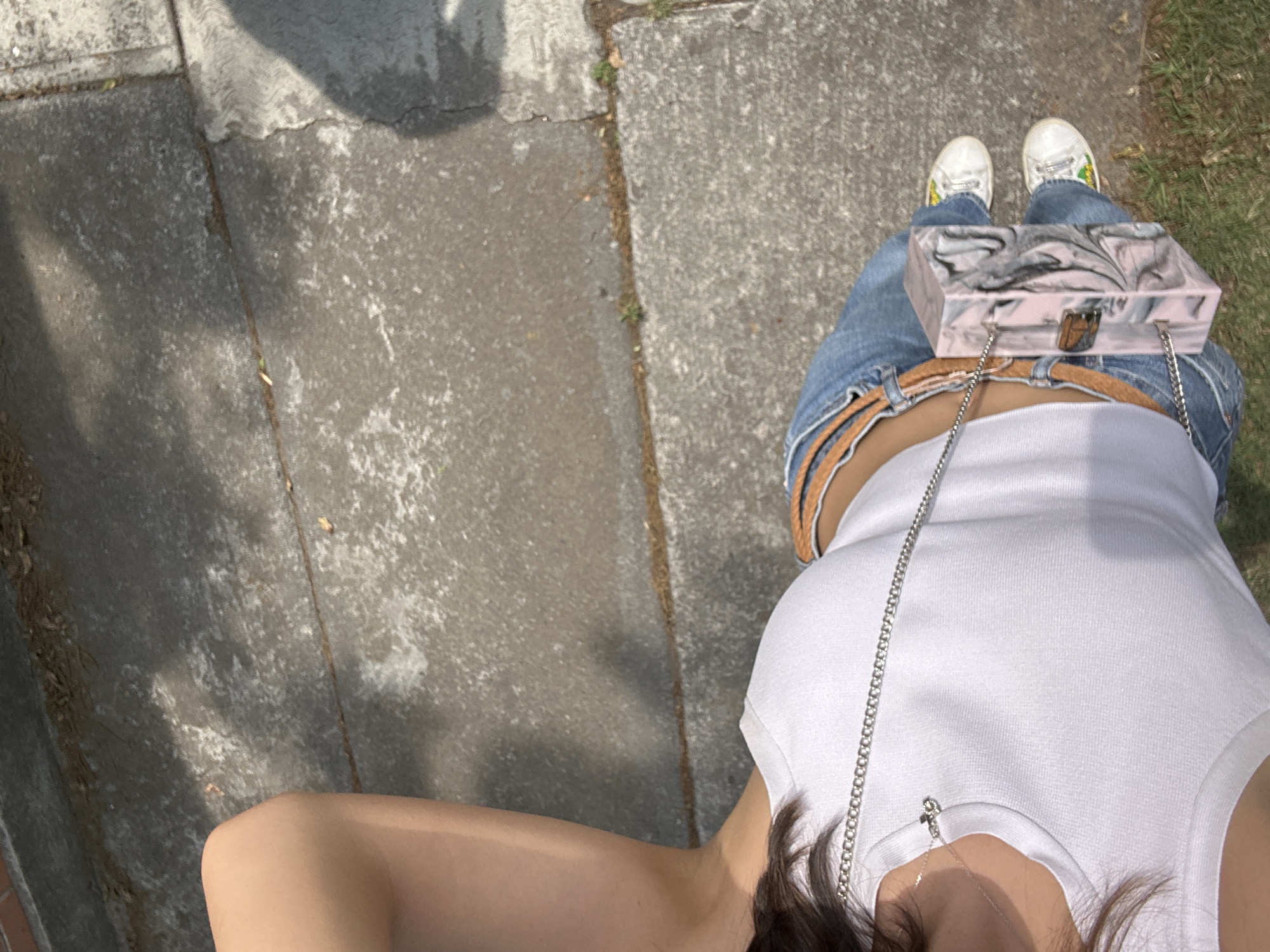 The image shows a person standing on a sidewalk next to a grassy area. The sidewalk is made of gray concrete slabs. The person is wearing light blue jeans, white sneakers with green and yellow details, a white tank top, and a brown belt. They are carrying a small, rectangular purse with a silver chain over their shoulder. The photo is taken from a high angle, looking down at the person.