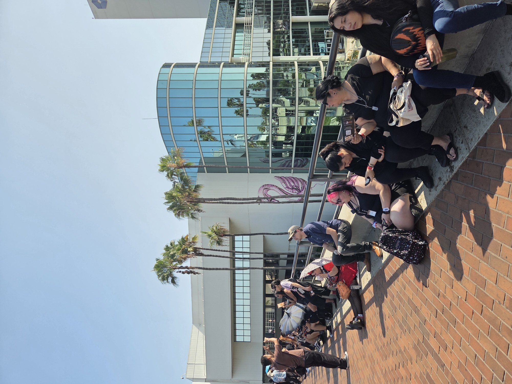 A group of people are sitting and standing in front of the Long Beach Convention Center in Long Beach, California. The convention center has a modern, curved glass facade that reflects the palm trees and clear blue sky. Several people are seated on a low wall, some holding phones or drinks, while others stand and converse or take pictures. A man in a brown shirt is taking a photo with his phone. Palm trees line the walkway, and the ground is paved with bricks. The overall scene suggests a warm, sunny day, possibly during an event at the convention center.