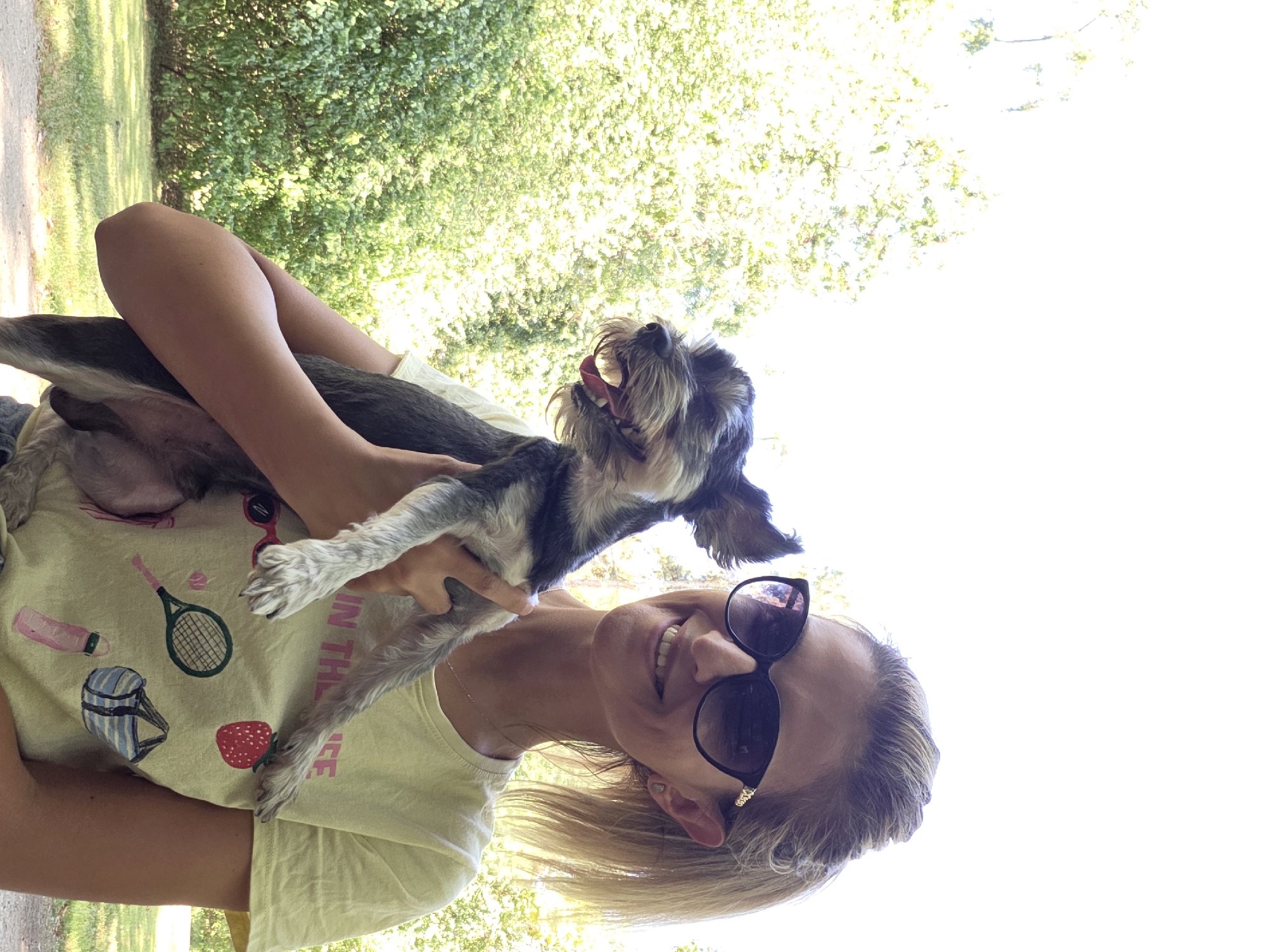 A young woman with blonde hair pulled back in a ponytail and wearing large dark sunglasses is holding a small grey and black dog in her arms. The dog is facing to the left and appears to be smiling, with its mouth open and tongue visible. The woman is wearing a light yellow t-shirt with various graphics printed on it, including a strawberry, a tennis racket, sunglasses, a water bottle, and a duffel bag. She is also wearing a necklace. The background consists of lush green trees and foliage, suggesting an outdoor setting, likely a park. The lighting is bright, indicating it is daytime. The scene is a portrait of the woman and her dog. There is no identifiable information in the image that places the scene in Munich, Germany.