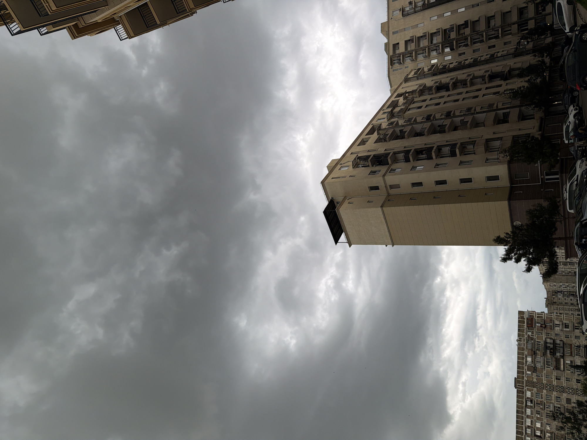 The image shows a low-angle view of several multi-story buildings in Tbilisi, Georgia, beneath a cloudy sky. The buildings have balconies and are different colors, with one taller one appearing beige. Cars are parked along the street to the right of one of the buildings. The sky is dominated by grey and white clouds, partially obscuring the sun. There are no people visible in the image.