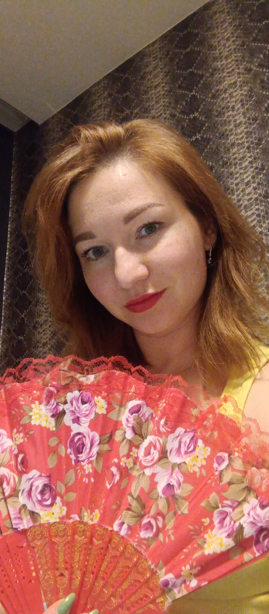 A close-up selfie of a red-haired woman holding a red fan with floral patterns. She has fair skin, freckles, green eyes, and is wearing red lipstick. An earring is visible in her left ear. The woman is wearing a yellow top. The fan is positioned in the foreground, partially obscuring her lower face and chest. The background is a textured, dark wall. The location is Unknown, Unknown.