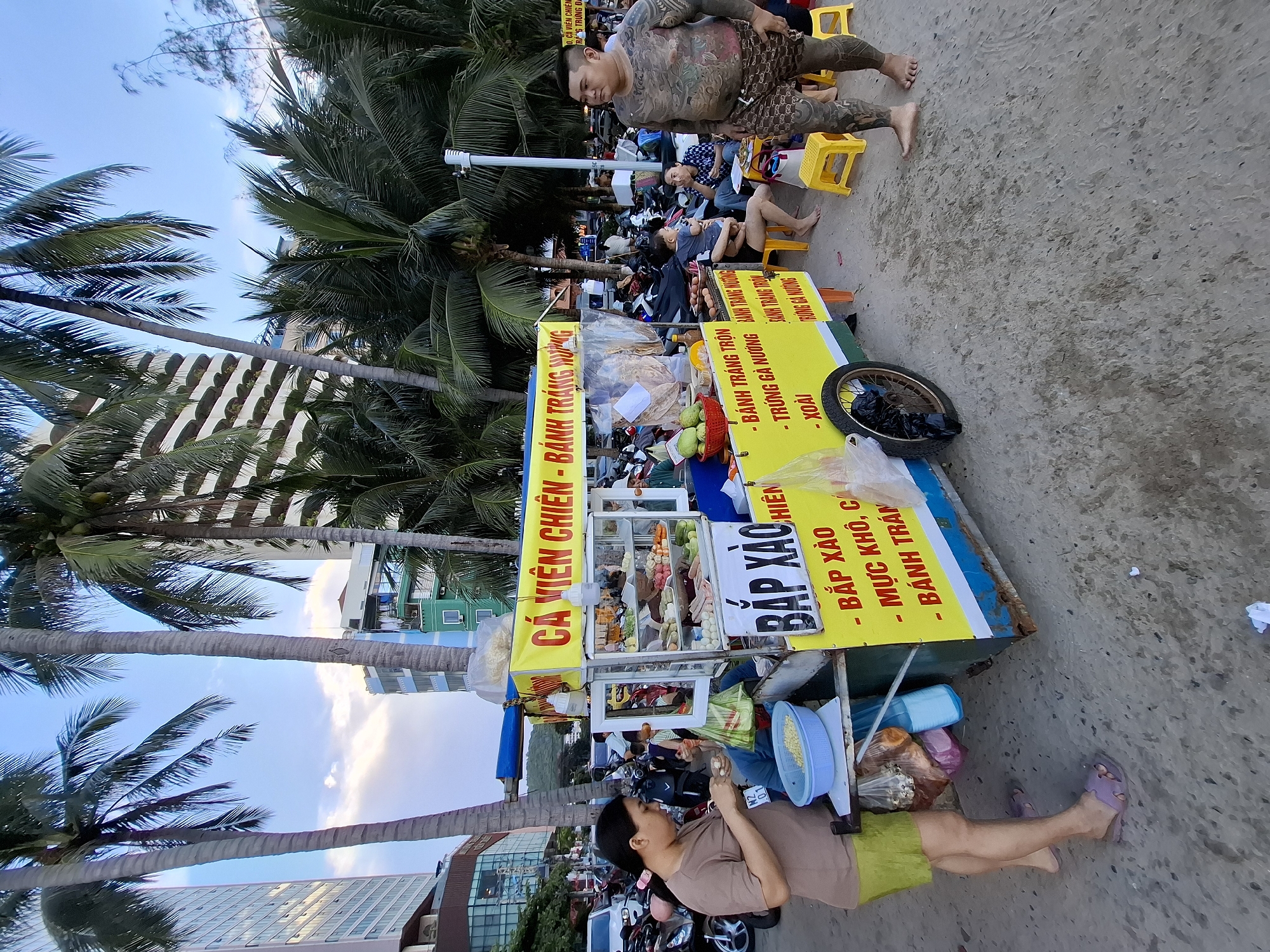 In the city of Nha Trang, Vietnam, a street food vendor operates a cart on a sandy beach. The cart, a brightly colored yellow and blue structure, displays a menu with Vietnamese text, advertising items like "Cá Viên Chiên" (fried fish balls), "Bắp Xào" (stir-fried corn), and "Trứng Gà Nướng" (grilled chicken eggs). Behind the cart, a busy street is visible with parked motorcycles and people. The background features tall buildings and lush palm trees, typical of a coastal city. A woman stands beside the cart, preparing food, while a heavily tattooed man stands in the foreground, observing the scene. The overall atmosphere is that of a lively beachside market.