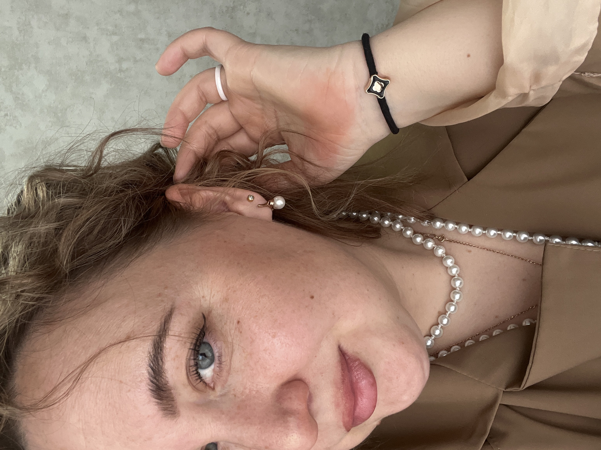 A close-up, sideways portrait features a person, likely female, from the chest up. The individual has light blue eyes, defined eyebrows, and pink lips, with visible freckles on their fair skin. Brown, wavy hair frames the face, with the person's right hand gently touching strands of their hair.
Several pieces of jewelry are worn: a simple white ring on the right ring finger, and a black cord bracelet with a small, black and gold stylized four-leaf clover charm on the right wrist. The right ear displays a small gold stud in the upper cartilage and a gold-toned earring with a pearl drop in the earlobe. Layered necklaces adorn the neck, including a prominent string of white pearls and delicate gold-colored chains.
The person is dressed in a brown or beige top, possibly satin-like, with visible pleating or draping. A lighter, cream-colored fabric is also visible on the right arm, suggesting part of the sleeve or an undergarment. The background is a soft-focus, light grey, textured surface. The lighting is even and soft, highlighting a calm or contemplative expression. No text is visible in the image.