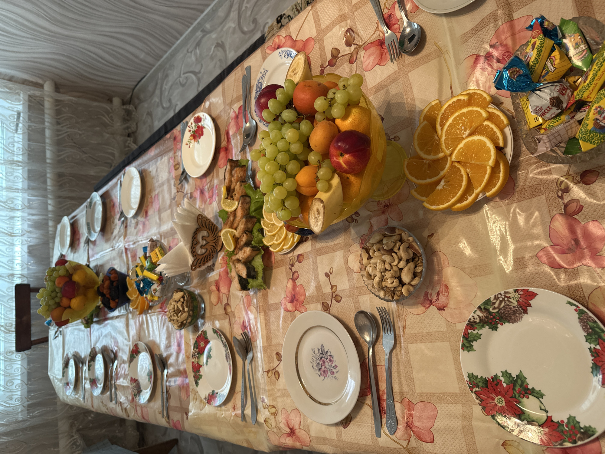 The image shows a table setting in what appears to be a home in Коноваловка, Russia. The table is covered with a floral patterned tablecloth and set with multiple plates, forks, and spoons. Various food items are arranged on the table including a bowl of fruit (grapes, oranges, peaches, bananas), sliced oranges, a dish of fish and lemons, a bowl of nuts, and a bowl of assorted candies. The setting suggests a gathering or celebration. There is no visible person in the frame. The interior of the building, presumably a house, is visible, including part of a window and curtains.