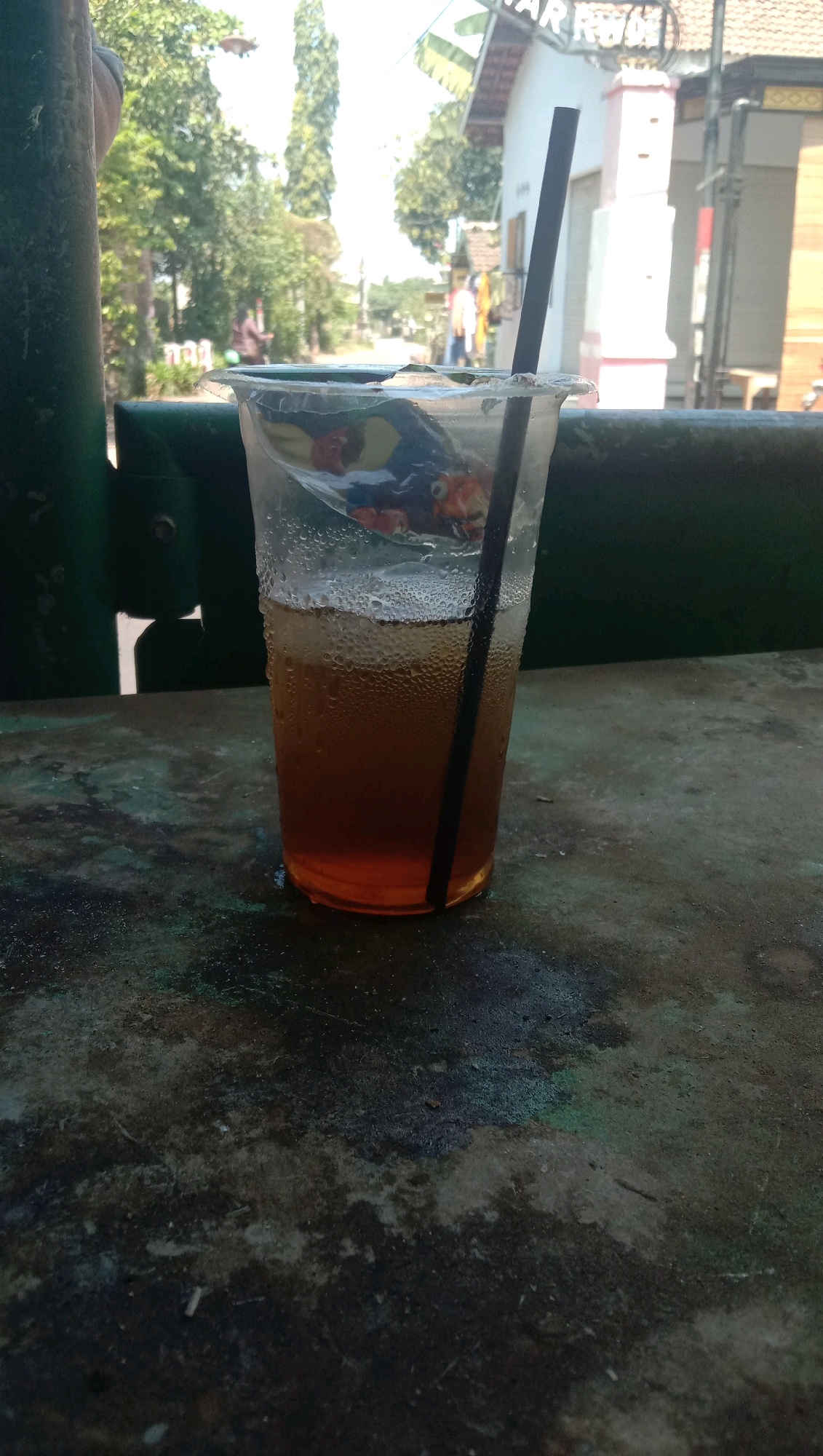A clear plastic cup with a black straw and iced tea is placed on a dark, textured surface, possibly a table. Condensation covers the outside of the cup, indicating the drink is cold. The cup has a colorful, possibly cartoonish, image on its inner lining. In the background, a blurred scene suggests an outdoor setting with trees, buildings, and a street. A sign that appears to read "KAR RWO" is visible on one of the buildings. The lighting suggests it is daytime, possibly a bright or sunny day. The image appears to be taken in the city of Karang Anyar, Indonesia, given the context provided. There are no people clearly visible interacting with the cup or in the immediate foreground. The overall scene is casual and depicts a beverage in an outdoor urban environment.