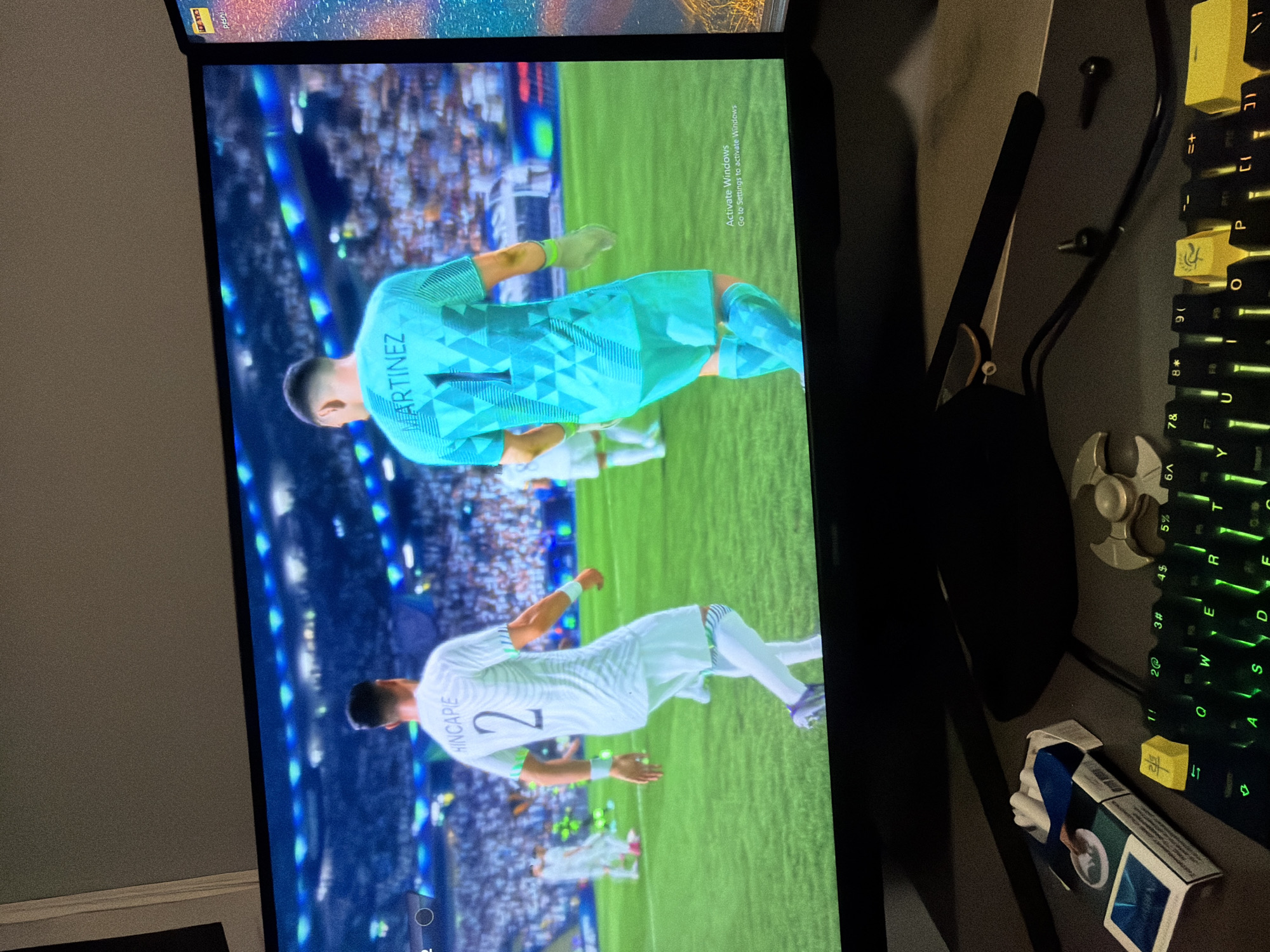 The image shows a screen displaying a FIFA video game, featuring soccer players Martinez (number 1, in turquoise) and Hincapie (number 2, in white). The environment appears to be a stadium with a crowd. The scene is located in a room with a keyboard (with yellow and green-lit keys), a stand, a cigarette box, a spinner and several screws. An "Activate Windows" watermark is visible on the screen.