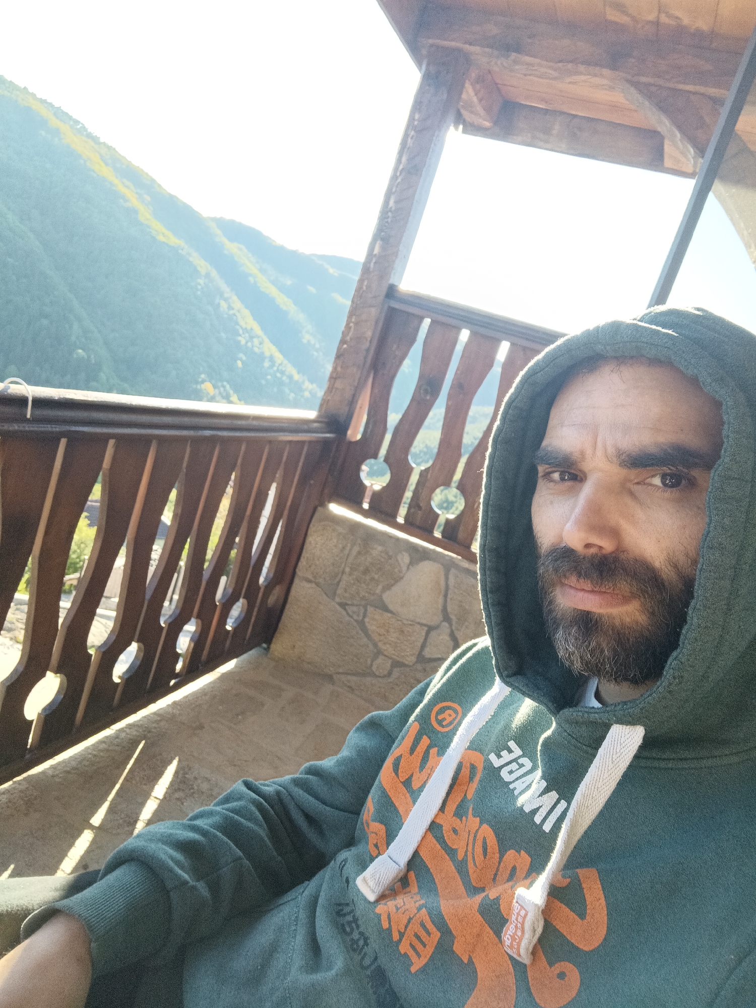 A man wearing a green hooded sweatshirt with white drawstrings is seated on a balcony with a wooden railing and a stone wall. Outside the balcony are mountains, likely in the vicinity of Vrata, Bulgaria. The man is facing the camera and has a beard. Sunlight casts shadows across the balcony.