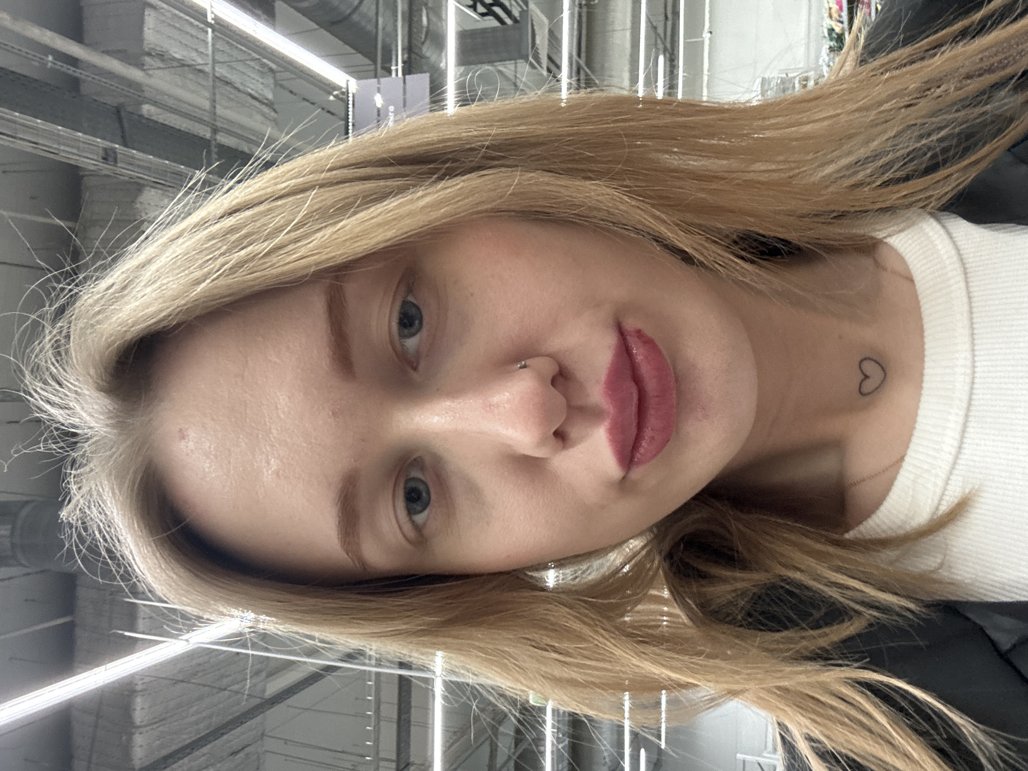 A female individual is depicted in a close-up, frontal portrait, positioned within an indoor commercial establishment in Rostov-on-Don, Russia. The subject has light blonde hair, which appears slightly dishevelled with flyaways, and light blue eyes. A small silver hoop nose ring is present on her left nostril. Her lips are coated with pinkish-red lipstick, with a noticeable smudge or wear mark on the lower lip. She is dressed in a white ribbed top, partially obscured by a dark-colored jacket or coat. A thin gold chain necklace is visible, and a small black outline heart tattoo is located on the left side of her neck, above the collarbone. The background reveals a high, industrial-style ceiling featuring exposed grey ductwork, pipes, and multiple linear fluorescent light fixtures arranged in rows. The environment suggests a large retail store, supermarket, or a similar commercial space with visible utilities. Distant, out-of-focus shelving or displays are faintly discernible in the background.