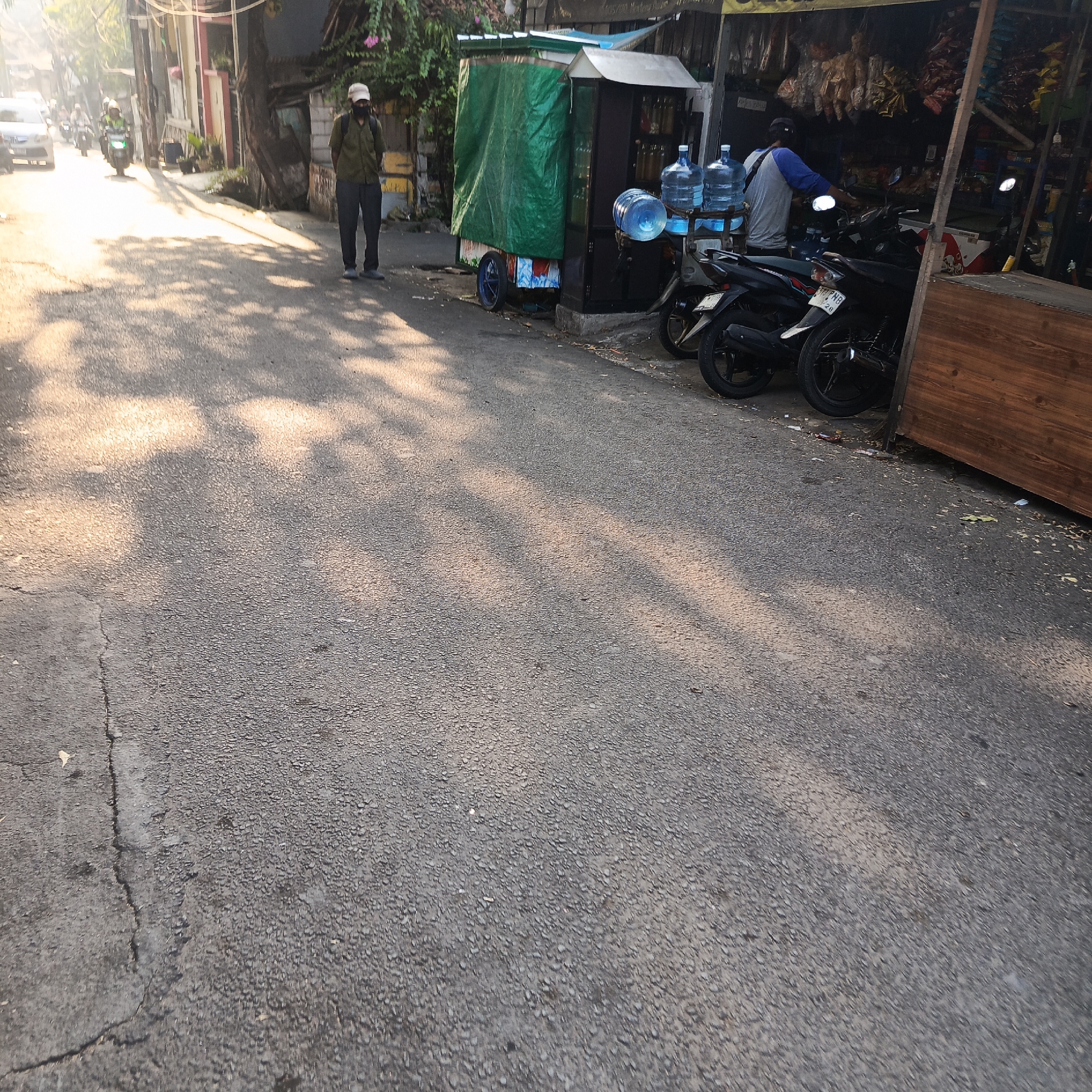 A street scene in South Jakarta, Indonesia, depicts a well-lit asphalt road with dappled sunlight casting shadows. A man wearing a cap and a dark face mask stands on the left side of the road, dressed in a olive green shirt and dark pants. To his right, a mobile cart is parked, covered with a green tarp and adorned with colorful decals. Two large water cooler jugs are visible on a rack attached to the cart. Further to the right, a stall is set up, displaying various items including bottled drinks and packaged goods. Two motorcycles are parked in front of the stall. The surrounding environment includes buildings and trees, with some of the buildings displaying a light color. The road appears to be a public thoroughfare with moderate traffic, as indicated by the presence of a car and several motorcycles in the background.