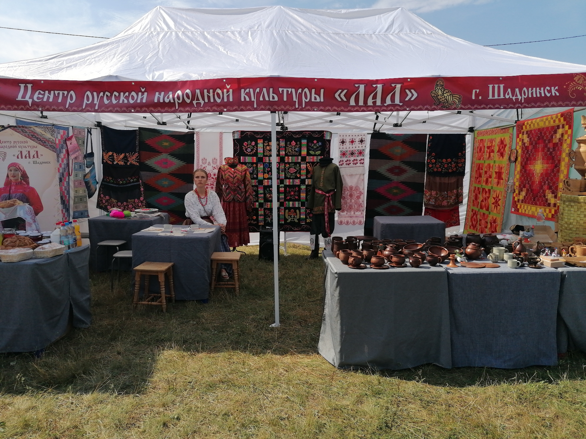 The image depicts an outdoor market stall under a white tent, likely set up for a cultural festival or event in Shadrinsk, Russia. A banner across the top of the tent reads "Центр русской народной культуры «Лада» г. Шадринск" (Center of Russian Folk Culture "Lada" Shadrinsk).
In the foreground, several tables are covered with dark grey tablecloths. The table on the left displays bread loaves and jars of preserves, along with bottles of liquid. A woman in traditional folk attire, wearing a white embroidered blouse and a red beaded necklace, sits behind this table. To her right, another table is adorned with various ceramic items, including cups, saucers, and pots. The background features traditional Russian textile art, such as embroidered cloth, woven rugs, and quilted wall hangings. Two mannequins are dressed in traditional garments. The ground is covered with dry grass, suggesting an outdoor setting.