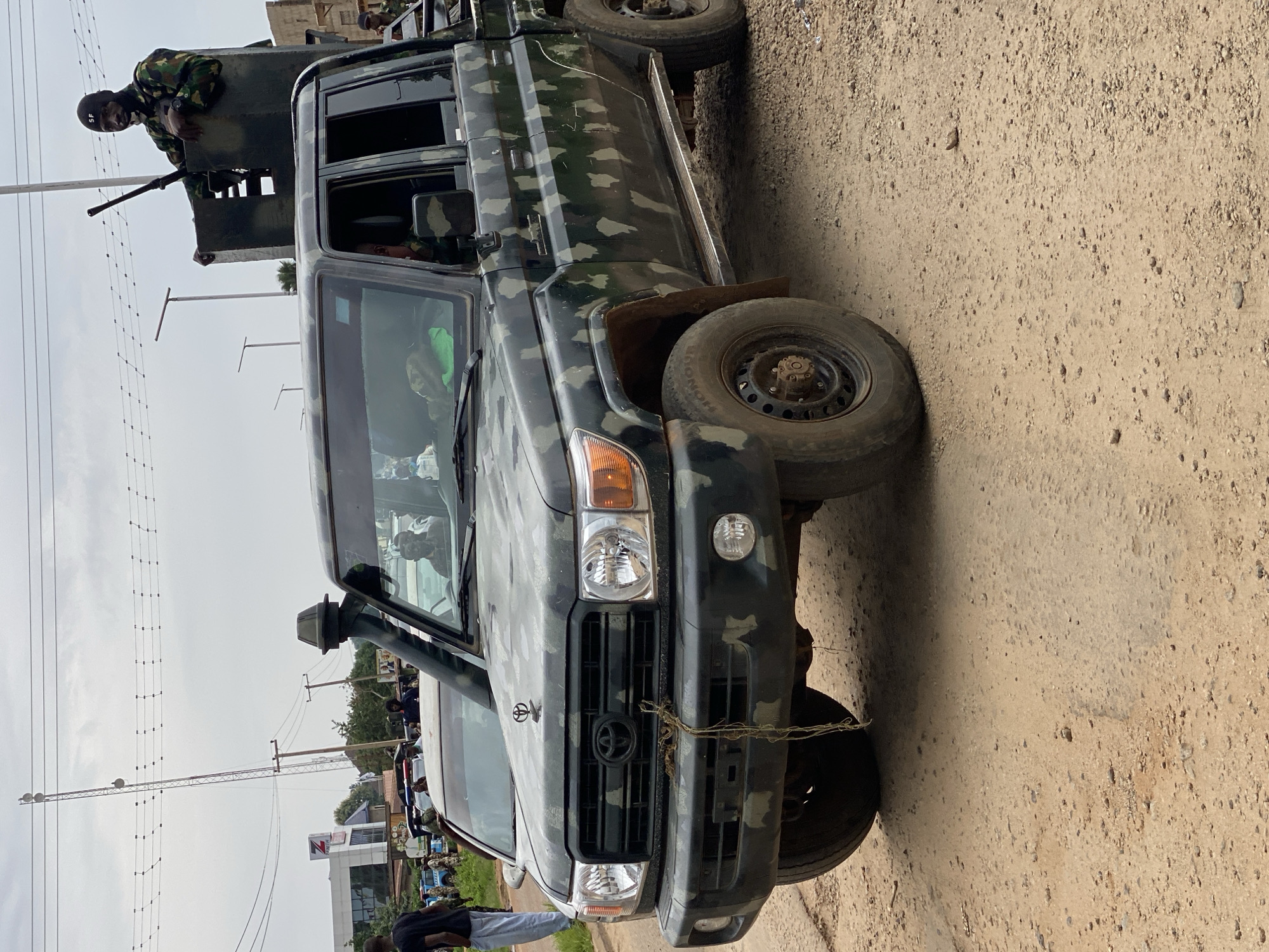 A military vehicle, camouflaged with a green and grey pattern, is parked on a dirt road. The vehicle has a gunner positioned in a turret on the back with a heavy weapon. The Toyota logo is visible on the front grill. Power lines and a building are in the background. People are visible in the distance. The location is Maigizo, Nigeria.