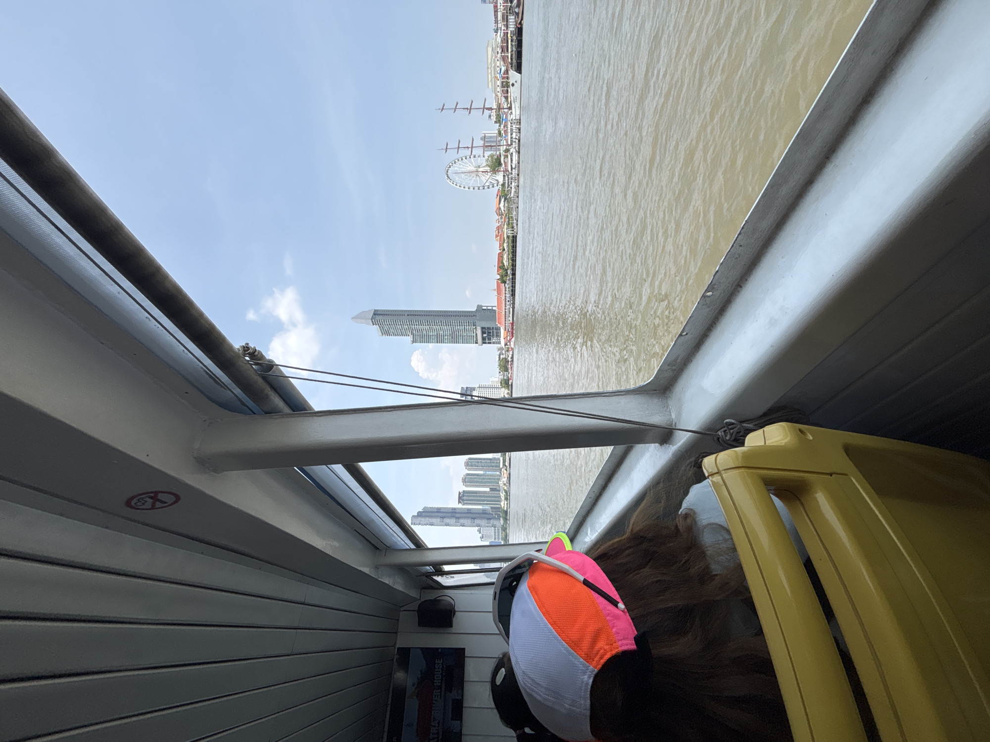 The image is taken from inside a boat on a river in Bangkok, Thailand, with a view of the cityscape. The brown water of the river fills the right side of the frame. In the background are various buildings, including a prominent skyscraper and a large Ferris wheel. The sky is blue with some scattered clouds. Inside the boat, the left side is lined with white, horizontally corrugated walls, with a "no smoking" symbol visible. A metal support structure crosses the view towards the center. In the foreground, a person with brown hair is wearing a colorful baseball cap, and part of a yellow object is visible.