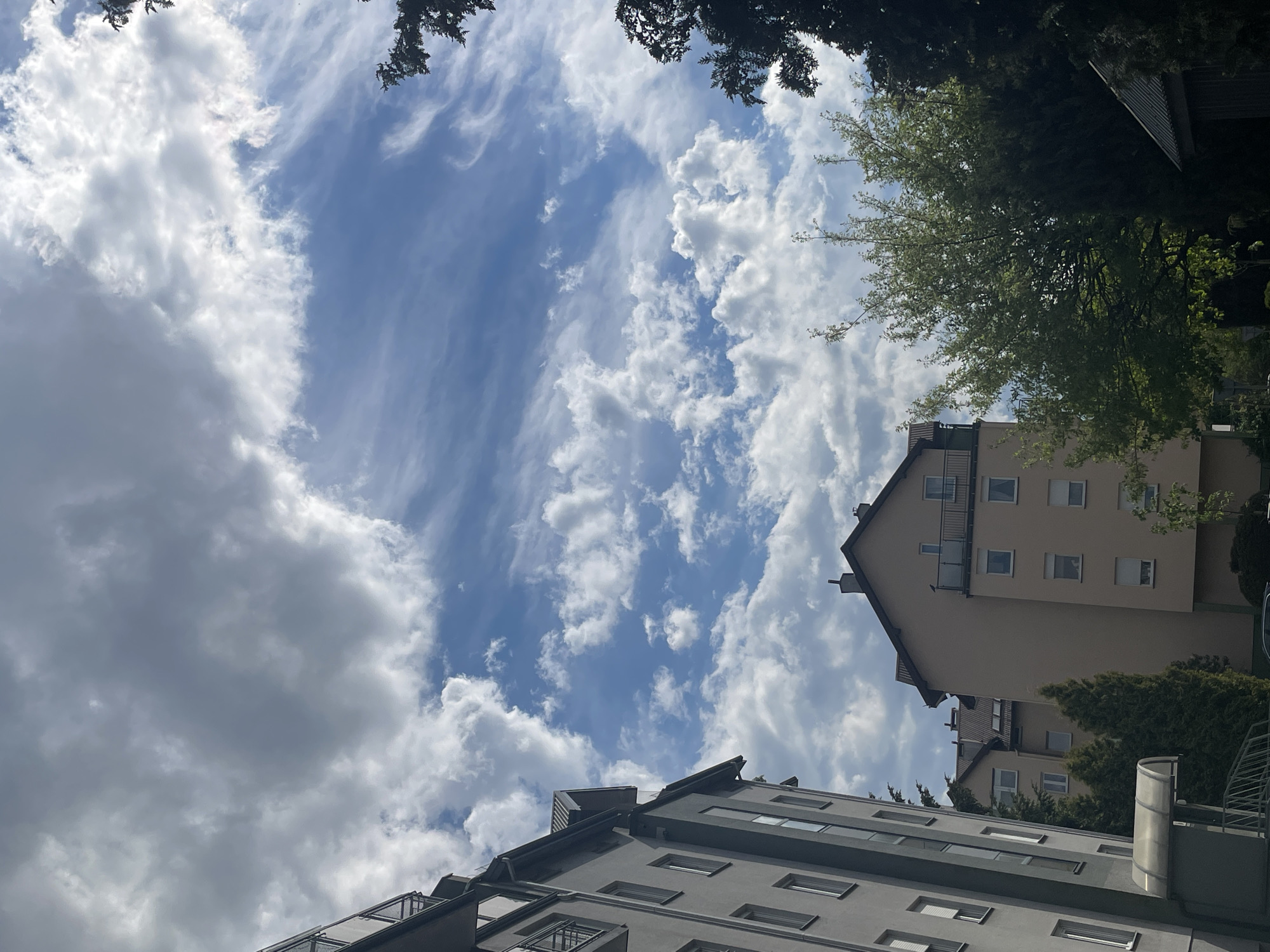 The image captures a low-angle view of several buildings against a cloudy sky. One building is light gray with dark gray trim, featuring multiple rectangular windows and small balconies. Another building is beige, with a similar design and gable roof. Patches of green trees are visible between and around the buildings. The sky is a mix of blue and white, with scattered cumulus clouds. The scene appears to be an urban residential area, though the specific location is unknown.