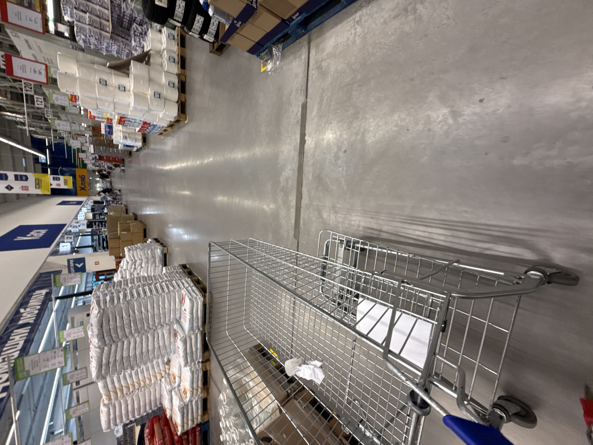 The image shows an interior view of a large retail store, possibly a warehouse or hypermarket, in Stara Zagora, Bulgaria. The perspective is from the point of view of a person pushing a shopping cart with a blue handle. Rows of merchandise are stacked on pallets on either side of a wide, concrete aisle. Visible products include toilet paper, paper towels, flour, and other packaged goods. The overall lighting appears to be bright and artificial.