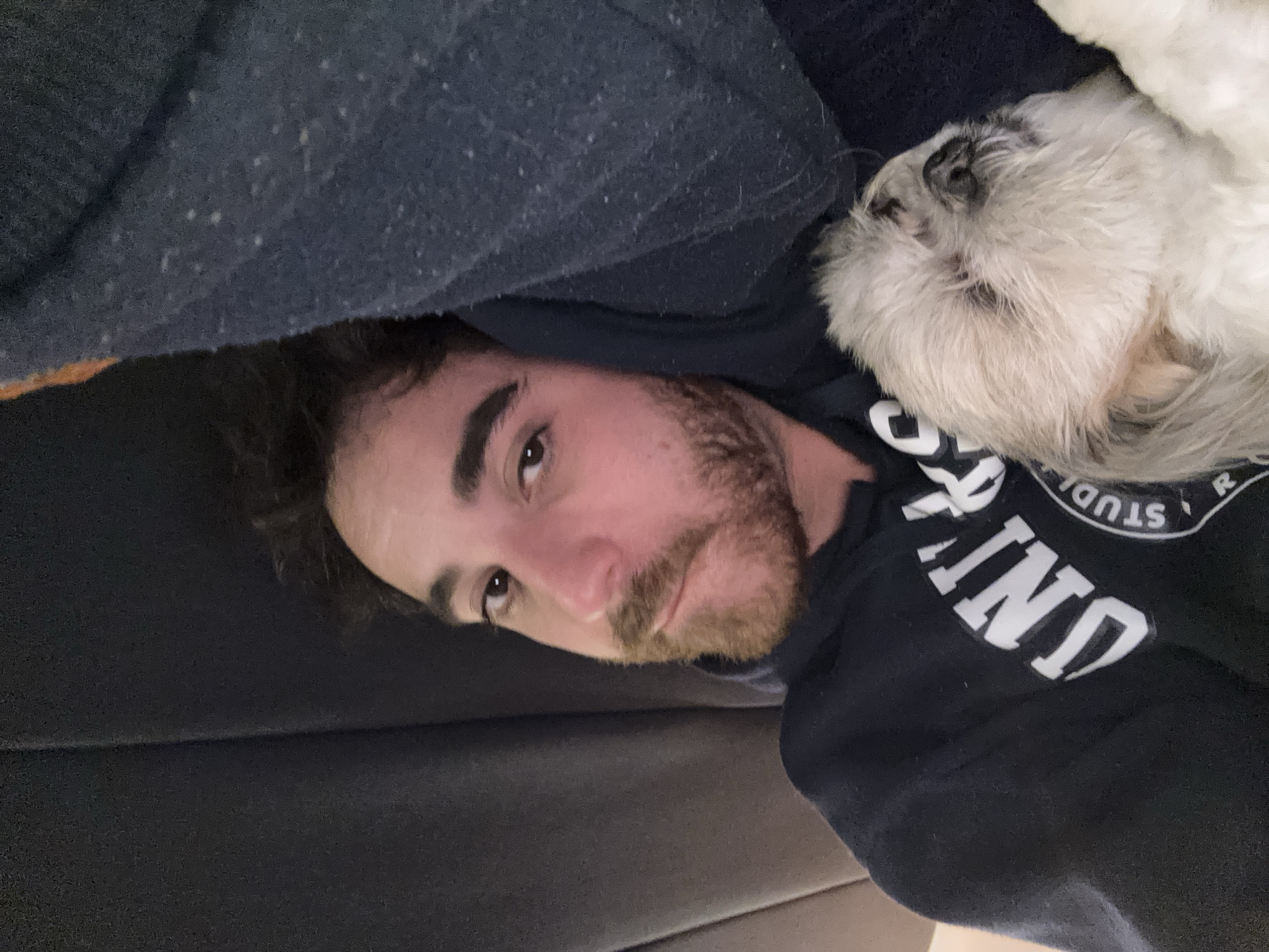 A man with brown hair and a beard is lying down on a dark gray surface, possibly a couch. A small white dog is resting on his right shoulder. The man is wearing a dark blue or black hooded sweatshirt with white lettering, partially obscured, but appearing to include "STUDIO" and "UNI". The background is a dark gray fabric. It is not possible to determine the location as Santa Clara Ocoyucan, Mexico from the visual information in the image.