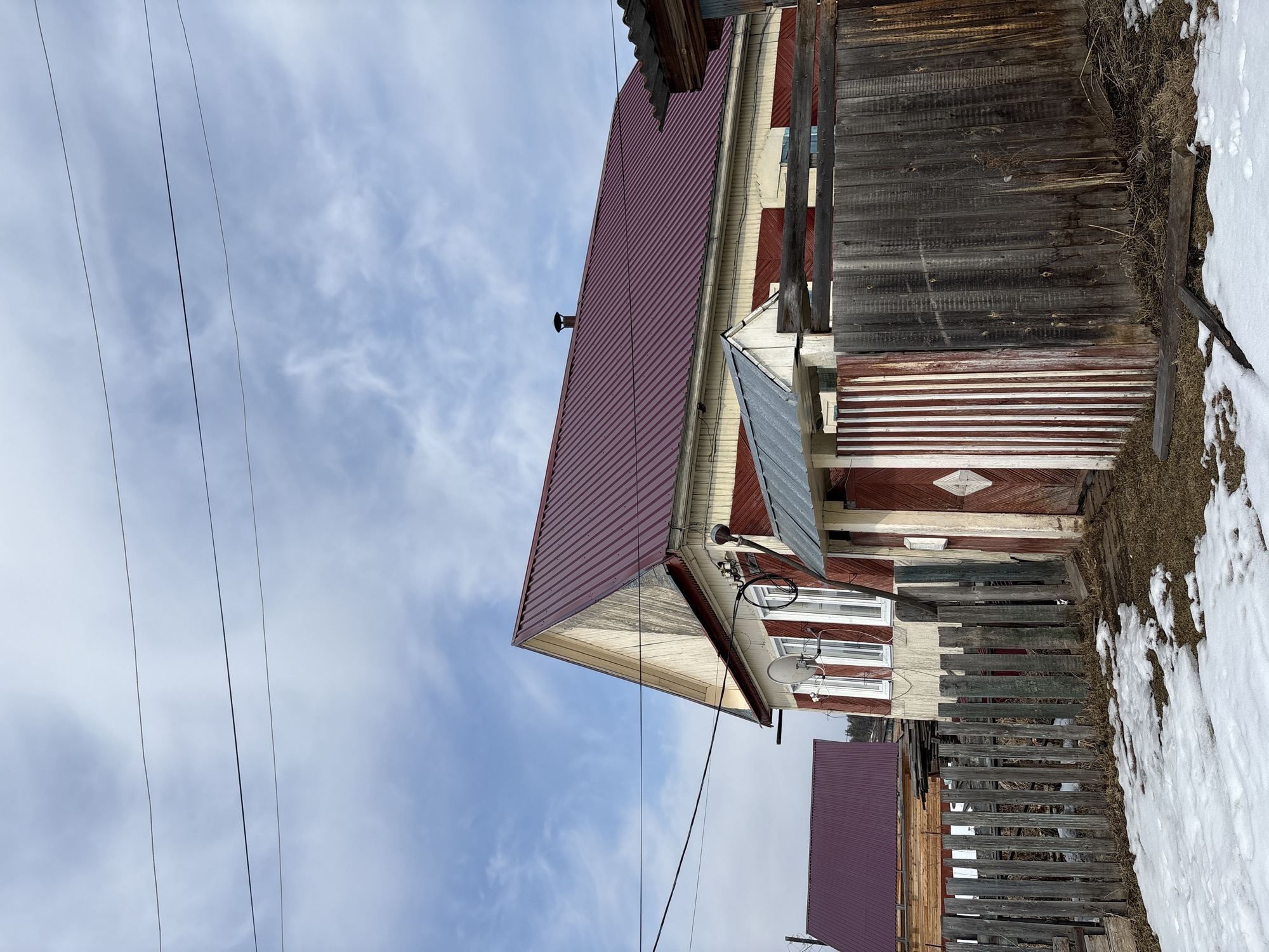The image depicts a weathered, two-story wooden house in Невон, Russia. The house has a red corrugated metal roof, and its walls are painted in alternating bands of cream and red. A satellite dish is mounted on the side of the house near the window. The fence appears to be made of aged wood. Patches of snow are visible on the ground near the fence and the side of the building. Electrical lines are visible in the foreground against a sky with scattered clouds.