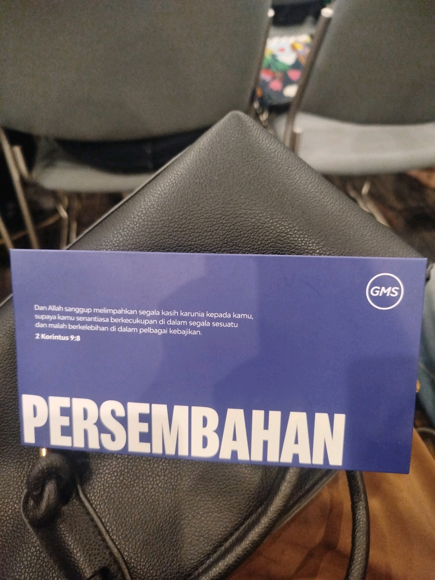 A blue card with white text lies on a black leather surface, possibly a bag. The card has a Bible verse and the word "PERSEMBAHAN" printed on it. In the upper right corner, a circular logo with "GMS" inside it is visible. The background shows blurred chairs and a hint of floral fabric. The image appears to be taken indoors, likely in a church or event hall in Sidoarjo, Indonesia. There are no people clearly visible in the frame.