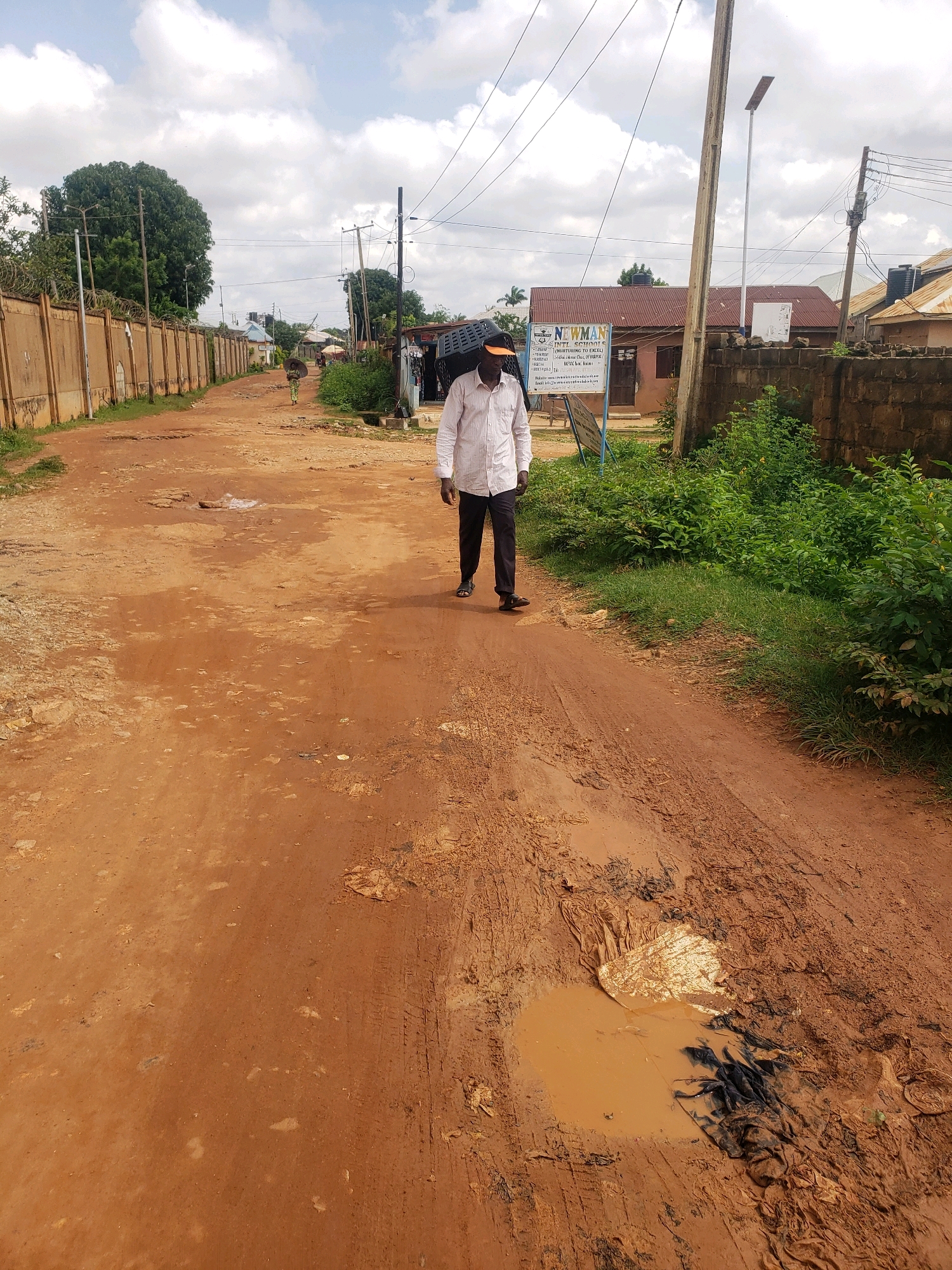 A man in a light-colored long-sleeved shirt and dark pants walks down a dirt road in Sabon Tasha, Nigeria. He is wearing an orange cap and black sandals. He is carrying a black object, possibly a basket or crate, on his left shoulder. The road is muddy and has several large potholes filled with water and debris, including what appears to be plastic bags and fabric. The surrounding area is lined with walls, vegetation, and buildings. A sign for "NEWMAN INT'L SCHOOLS" is visible on the right side of the road, mounted on a pole next to a building with a corrugated iron roof. Overhead power lines are strung across the scene, supported by utility poles. The sky is cloudy.