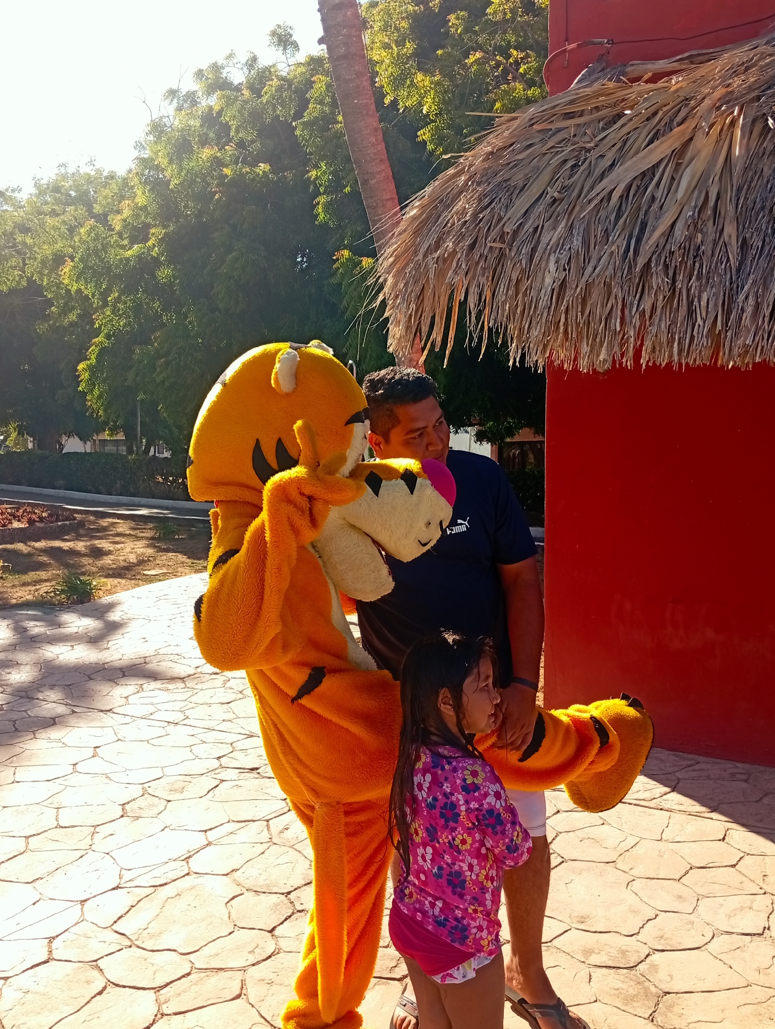 A man, a child, and a person in a tiger mascot costume are standing on a stone-paved area. The man is wearing a dark blue t-shirt, and the child is wearing a pink floral swimsuit cover-up. The tiger mascot is predominantly orange with black stripes. They are positioned next to a red structure with a thatched roof. In the background, there are green trees and a palm tree. The scene is illuminated by sunlight, casting shadows on the ground. The location appears to be Playa El Agua, Venezuela.