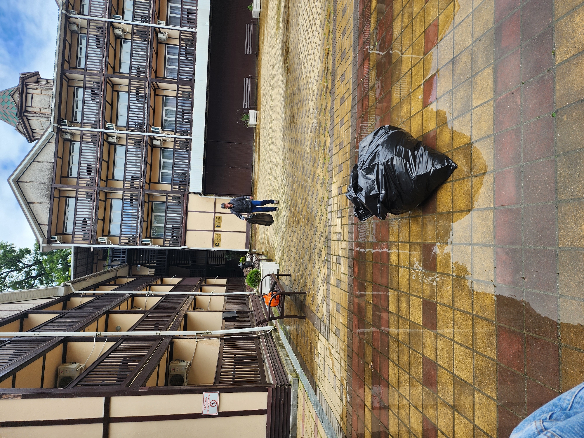 The image depicts an outdoor scene in Sochi, Russia. Two multistory buildings, characterized by balconies with brown lattice railings and cream-colored walls, frame a rectangular courtyard paved with wet, yellow and brown checkered tiles. A man wearing a dark jacket and jeans walks away from the camera, carrying a black bag. A large black trash bag sits on the wet tiles near the right edge of the frame. A row of park benches, some with decorative plants in planters, runs between the buildings. The sky is partially visible and cloudy. A small sign with a crossed-out insect is visible on the bottom left building.
