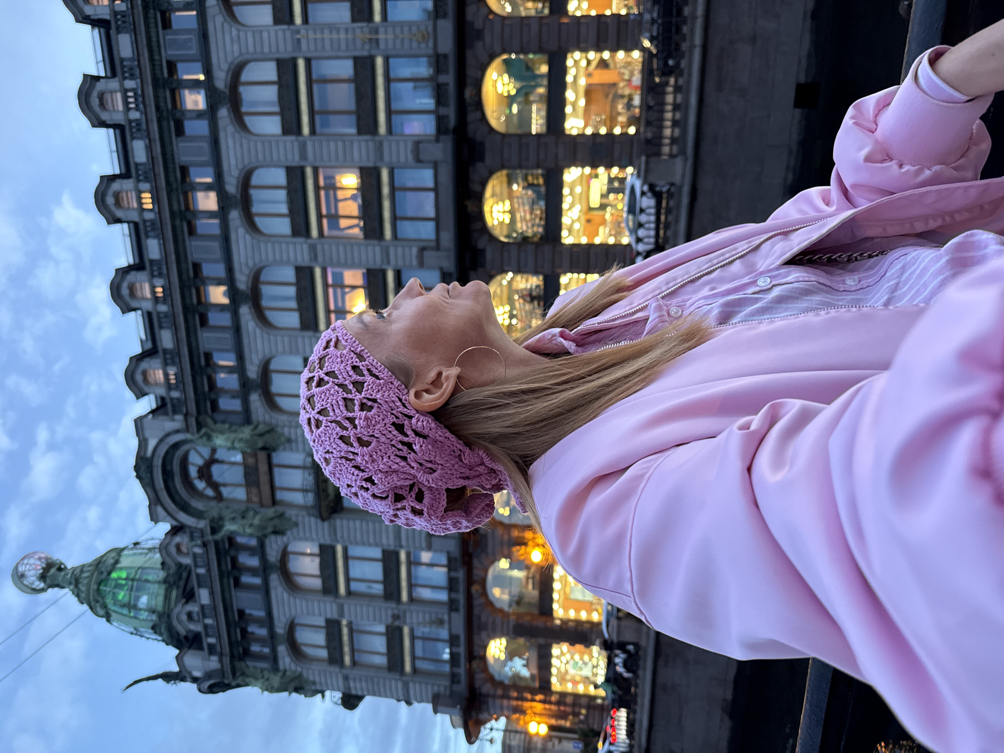 A woman wearing a pink knitted hat and a pink bomber jacket is standing by a canal in Saint Petersburg, Russia. She is looking to the right of the frame, with the Singer House building in the background. The Singer House is a historic Art Nouveau building with a distinctive glass dome and a globe on top. The building is illuminated with lights, reflecting in the canal. The sky is overcast and a bit cloudy, suggesting it is dusk or early evening. The woman has blonde hair and is wearing a striped shirt underneath her jacket. She has a hoop earring in her visible ear. The scene is captured from a slightly low angle, with the canal railing in the foreground.