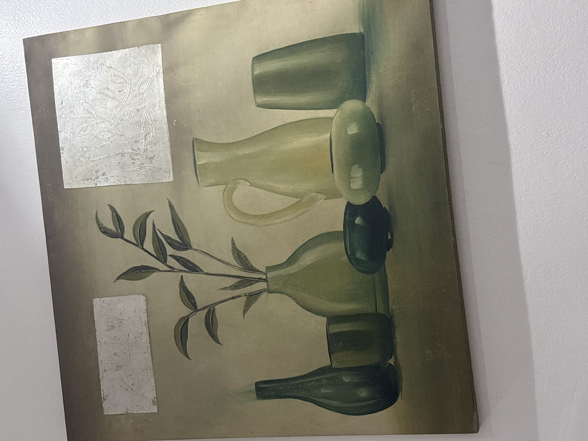 The image displays a still life painting, captured from a slightly angled perspective, appearing rotated 90 degrees counter-clockwise. The painting features an arrangement of various vessels, predominantly in shades of muted green, beige, and olive.
**Subjects:** The central subjects of the painting are several bottles, jugs, and cups, rendered in a stylized, somewhat abstract manner. A branch with dark green leaves emerges from one of the vessels, suggesting a plant or foliage element. In the top-left and bottom-left quadrants of the painting, there are two rectangular patches with a silver-grey, textured appearance, adorned with embossed or raised floral and vine patterns. The background of the painting is a soft, earthy gradient transitioning from lighter beige to darker olive tones.
**Activity or event taking place:** The painting depicts a static still life composition; no activity or event is taking place within the artwork itself.
**Notable details:** The overall color palette is subdued, evoking a natural, earthy feel. The objects within the painting cast subtle shadows, indicating a soft light source. The silver-grey textured patches provide a contrasting visual and tactile element against the painted vessels. The photograph shows the painting leaning against a plain white wall, which has a lightly textured surface. The shadow of the painting is visible on the wall, suggesting indoor lighting. The image captures the edge of the painting's frame on the right side.
**Visible text:** There is no visible text within the painting or on its frame.
