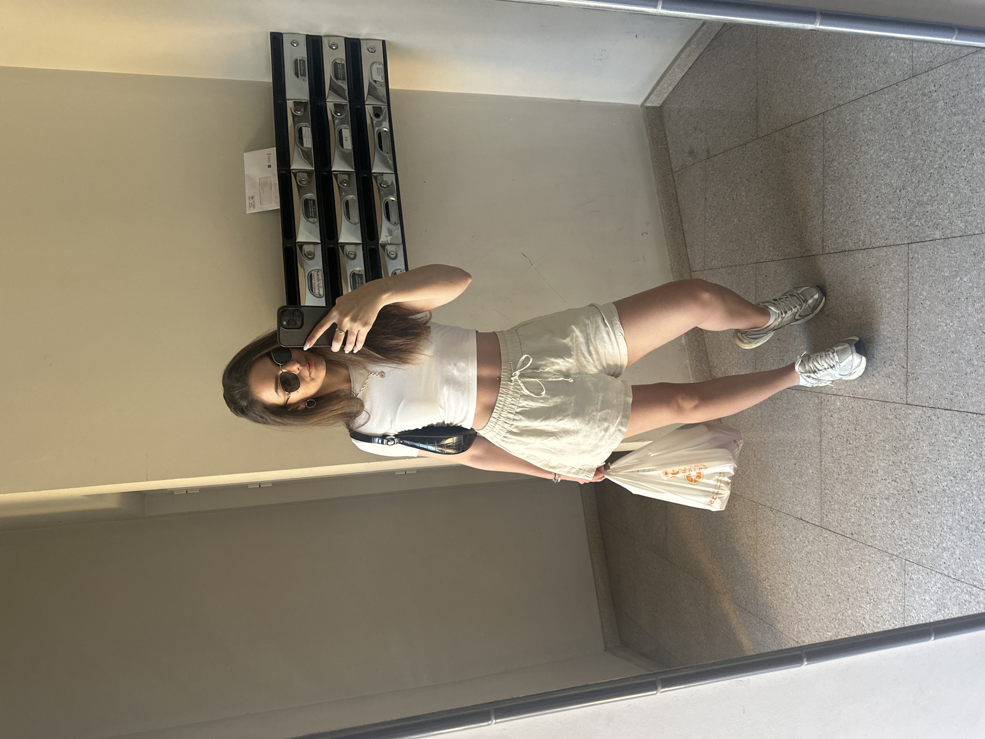 A woman is taking a selfie in front of a mirror. She is wearing sunglasses, a white crop top, and beige shorts. She has a black purse over her shoulder and is holding a white plastic bag. Behind her is a wall with mailboxes, and the floor is tiled. The location is not discernible from the image; therefore, it's impossible to tell that it is in Beniflà, Spain.