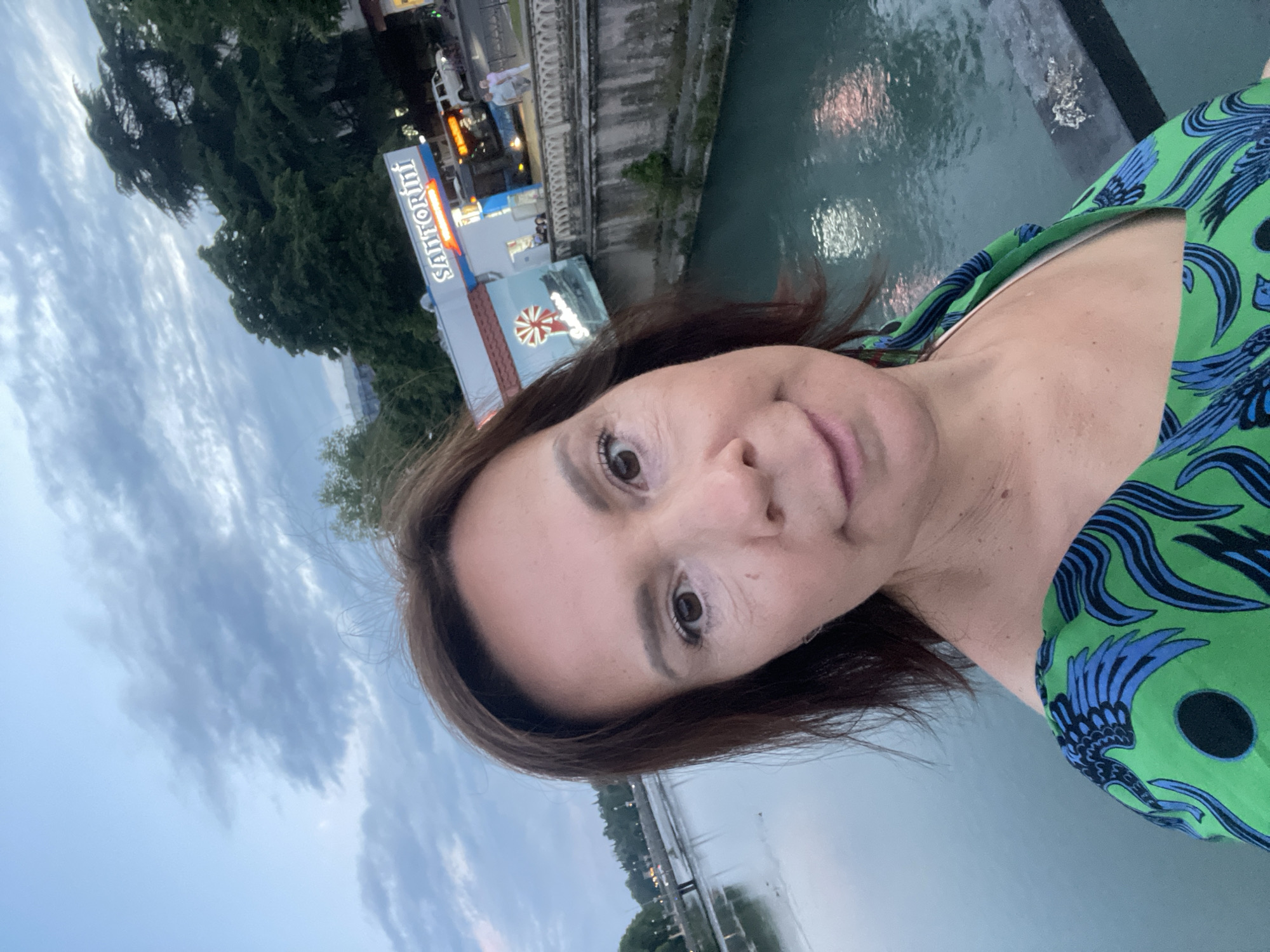 A woman with brown hair and brown eyes takes a selfie in front of a body of water, likely a river, in Sochi, Russia. She is wearing a green dress with a blue and black pattern. Behind her, a building with a sign reading "SANTORINI" is visible, along with trees and a cloudy sky. The water reflects the lights from the surrounding area, creating a shimmering effect. A bridge can be seen in the distance over the water.