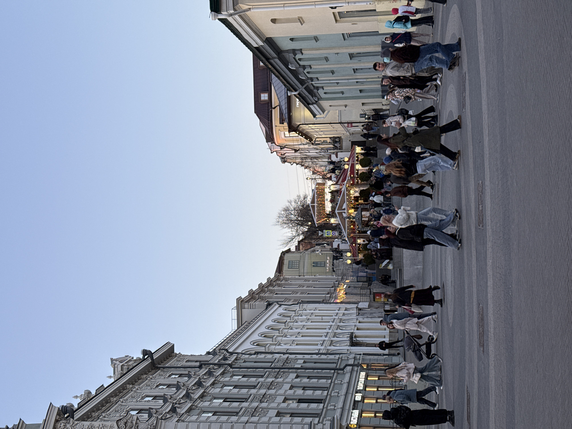A street scene in Kazan, Russia, shows a broad pedestrian walkway lined with buildings on both sides. The buildings vary in architectural style, some with intricate facades and others with simpler designs. The walkway is filled with people strolling and interacting. The sky is a clear light blue, and the scene suggests a lively urban environment with commercial activity indicated by store signage. Decorations are strung across the street.