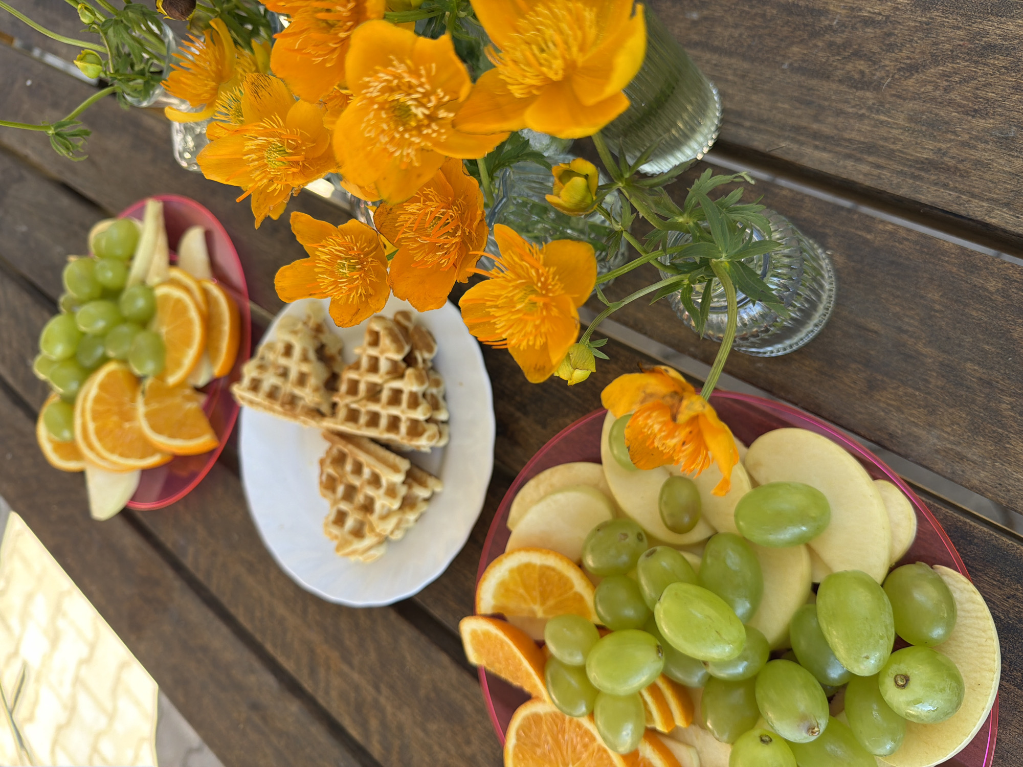 The image depicts a wooden table outdoors, with several plates of food and flowers. One plate contains sliced oranges, green grapes, and apple slices. Another plate holds waffles. In the foreground, a bouquet of vibrant orange flowers in small glass vases is arranged. The table surface is made of dark wood planks. The background is blurred, but appears to be an outdoor setting with patterned paving stones visible in the upper left corner. There are no people or animals present. The lighting suggests it is daytime.