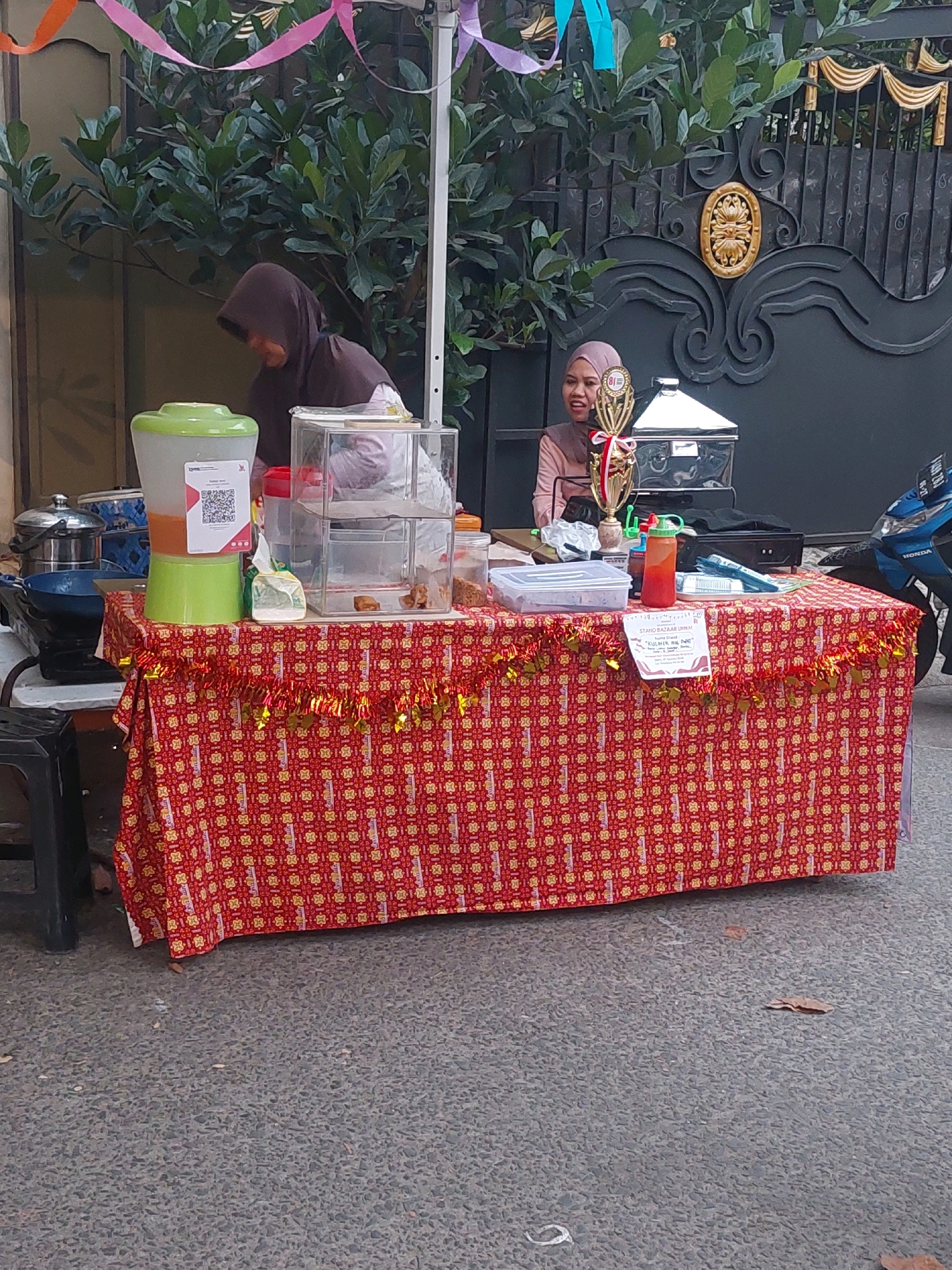 The image depicts a street-side stall in Depok, Indonesia, likely set up for a community event or bazaar. The stall is covered with a vibrant red tablecloth adorned with a repeating yellow floral pattern and a festive fringe of gold and red tinsel. On the table, a large green beverage dispenser with a QR code is visible, alongside a transparent display case filled with food items. To the right, a young woman wearing a pink hijab smiles and looks towards the camera, standing behind a trophy and a metallic container. Another person, partially obscured by a dark hijab, is also behind the stall. The background features lush green foliage and an ornate black metal gate, suggesting the stall is situated near a residential or commercial property. The ground is paved with asphalt, and a blue motorcycle is parked to the right of the stall. The overall scene suggests a lively outdoor market atmosphere.
