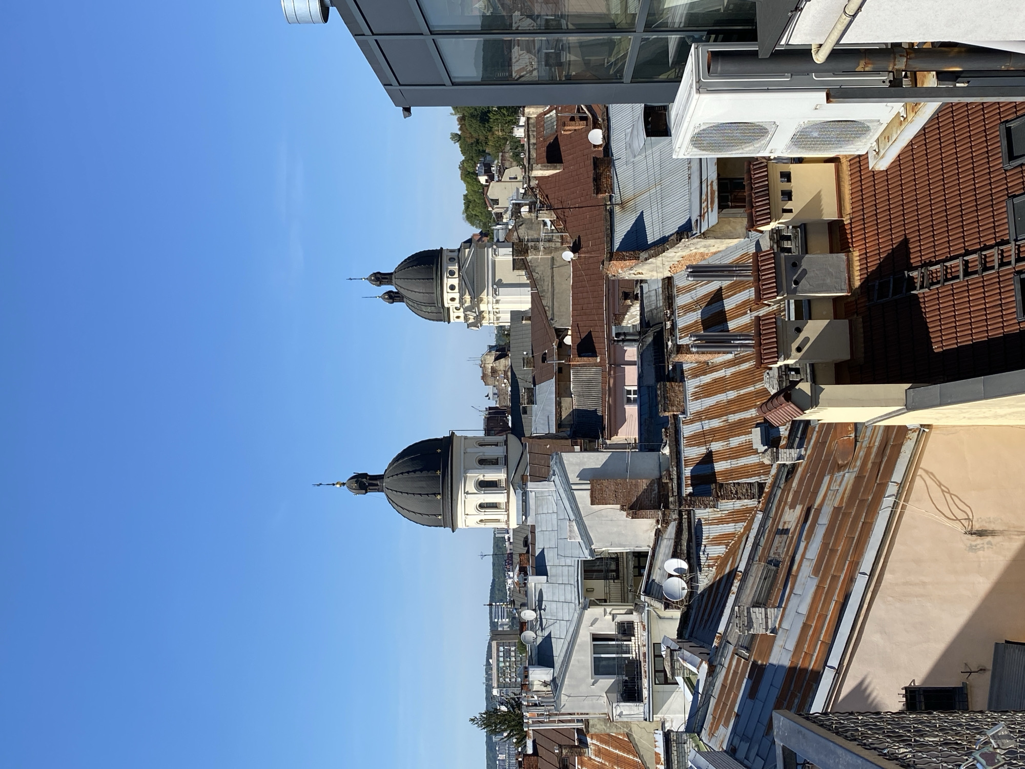 The image is an aerial view of the rooftops of Lviv, Ukraine, on a clear, sunny day. In the mid-ground, two prominent church domes with black coverings and gold crosses are visible against a bright blue sky. These domes belong to the Armenian Cathedral and the Dominican Church. The foreground and mid-ground are filled with a dense collection of buildings with varied roofing materials, including rusted metal, terracotta tiles, and gray sheeting. Chimneys of different shapes and sizes punctuate the roofscape. In the lower right corner, modern elements are present, including a glass-fronted building and an outdoor air conditioning unit mounted on a metal platform. The scene depicts an urban environment with a mix of historical and modern architecture, bathed in natural sunlight, casting shadows across the rooftops.