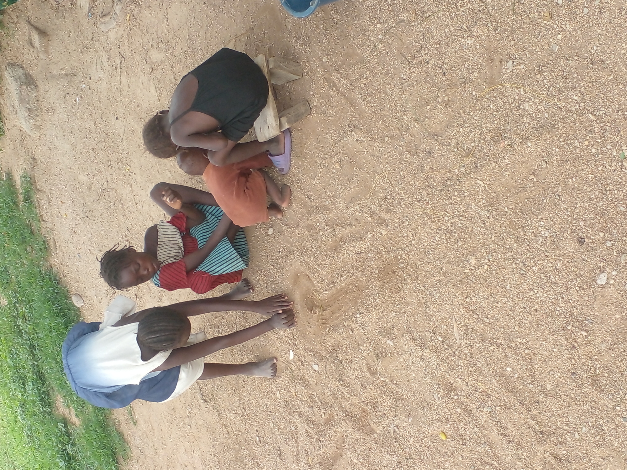 In an outdoor, sandy environment with sparse green grass in the background, four young individuals are present. One person, wearing a blue and white ombre dress, is bent over and appears to be drawing or playing in the sand with their hands. Another individual, in a red and striped top and a blue and green striped skirt, is sitting and looking down. A third person, in an orange garment, is kneeling and looking towards the ground. The fourth individual is seated on a small wooden stool, wearing a black tank top and shorts, and appears to be observing the others. The ground is composed of fine sand with small pebbles. The overall scene depicts children engaged in an activity outdoors.