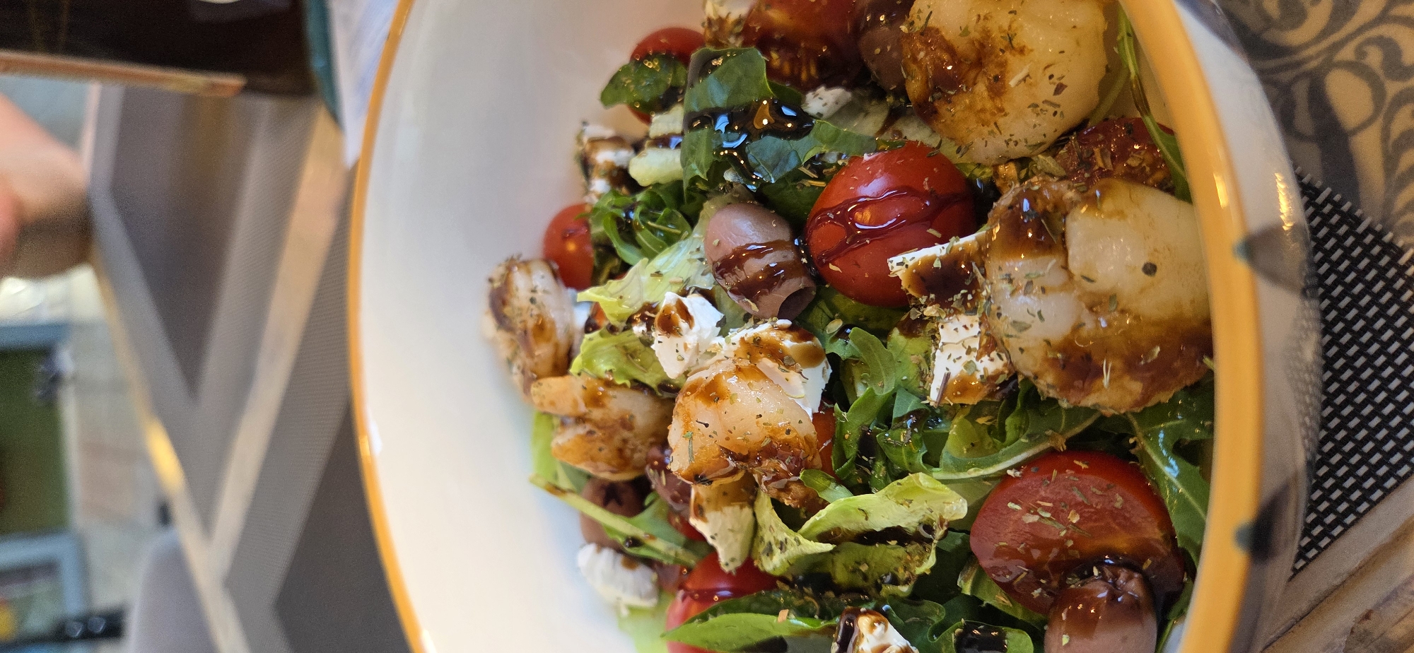 The image, captured in Lipetsk, Russia, depicts a close-up, high-angle view of a colorful salad served in a white bowl with an orange rim. The salad is composed of various ingredients including fresh green lettuce and arugula leaves, halved red cherry tomatoes, white cubes resembling feta cheese, dark brown olives, and several pieces of grilled or seared seafood, possibly scallops or large shrimp, which are golden-brown and appear seasoned with dried herbs. A dark, viscous balsamic glaze or similar dressing is generously drizzled over the entire salad. The background is out of focus, revealing a dining setting with a table partially covered by placemats featuring geometric patterns, one with dark and light grey stripes, and another with a black and white checkered design. A blurry human arm/hand is visible in the upper right background, and a dark bottle stands upright on the table to the right. The overall environment suggests an indoor dining area, likely a restaurant or cafe.