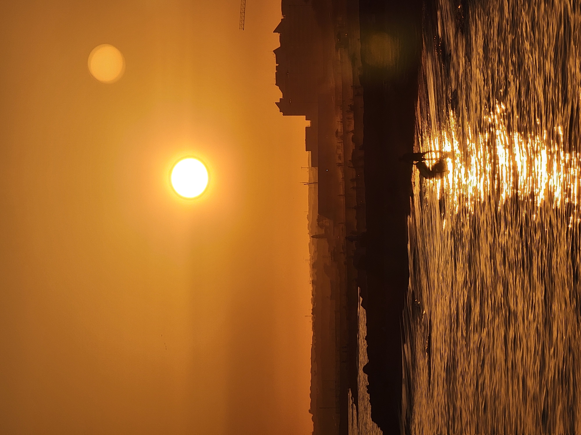 The image shows a sunset over the coast of Quarteira, Portugal. Two suns are visible in the sky, one larger and brighter than the other. The larger sun is setting over the horizon, casting a warm glow on the water and the surrounding buildings. The smaller sun is higher in the sky, appearing as a faint circle.
In the foreground, the ocean waves reflect the golden light of the sunset. A dark silhouette of a person is visible in the water, seemingly engaged in an activity such as paddleboarding or kayaking. The shoreline is lined with a rocky breakwater, beyond which a city skyline is visible, silhouetted against the bright sky. Numerous structures, including buildings and possibly cranes, can be discerned in the distance, indicating an urban environment. Small figures of people are scattered along the beach area between the breakwater and the city. A few small dark specks, possibly birds, are present in the upper left portion of the sky.