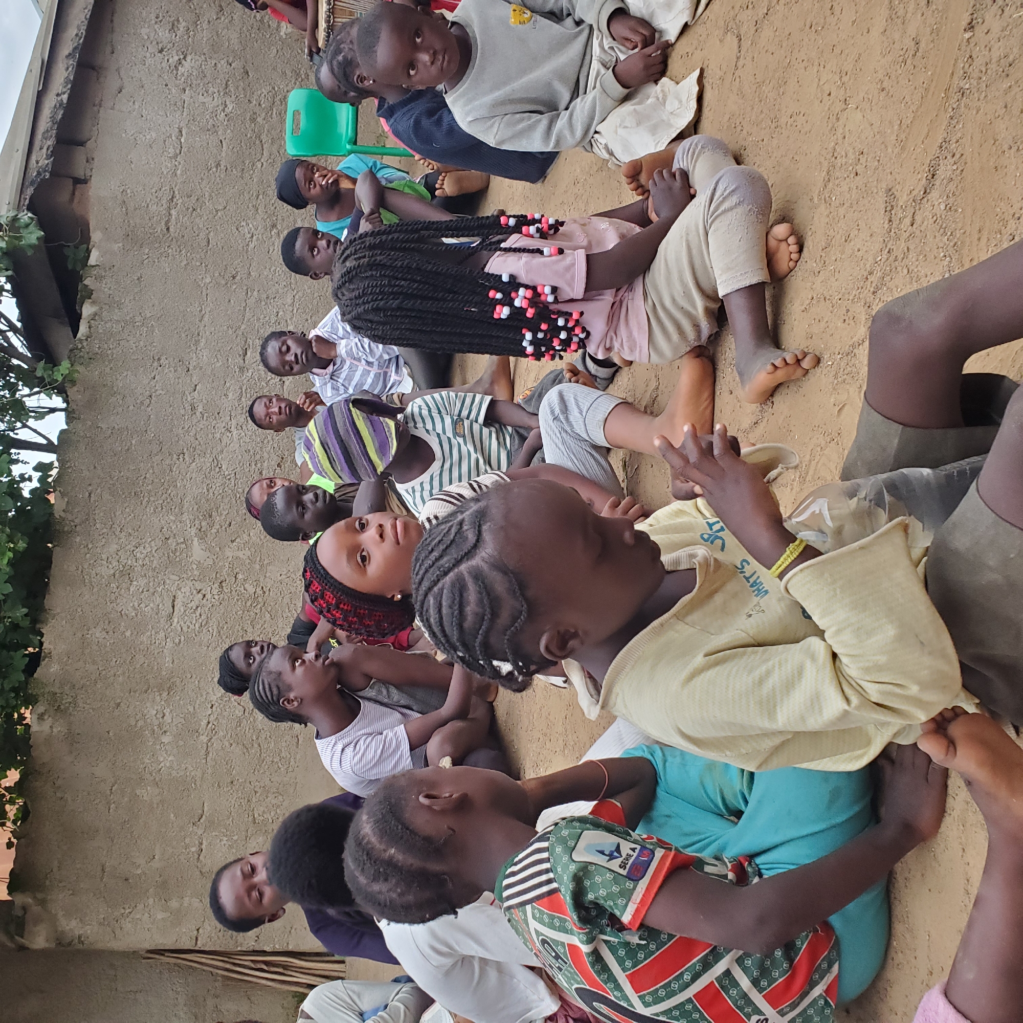 A group of children, appearing to be of African descent, are sitting on the ground in front of a textured concrete wall. They are dressed in casual clothing and some have head coverings. The children are facing in various directions, with some looking towards the viewer and others looking away. One child in the foreground is wearing a patterned shirt and has their hand near their face. Another child is wearing a yellow shirt and is holding a clear plastic bag. In the background, a green plastic chair is visible, and some foliage hangs from above the wall. The ground is composed of dirt and small pebbles.