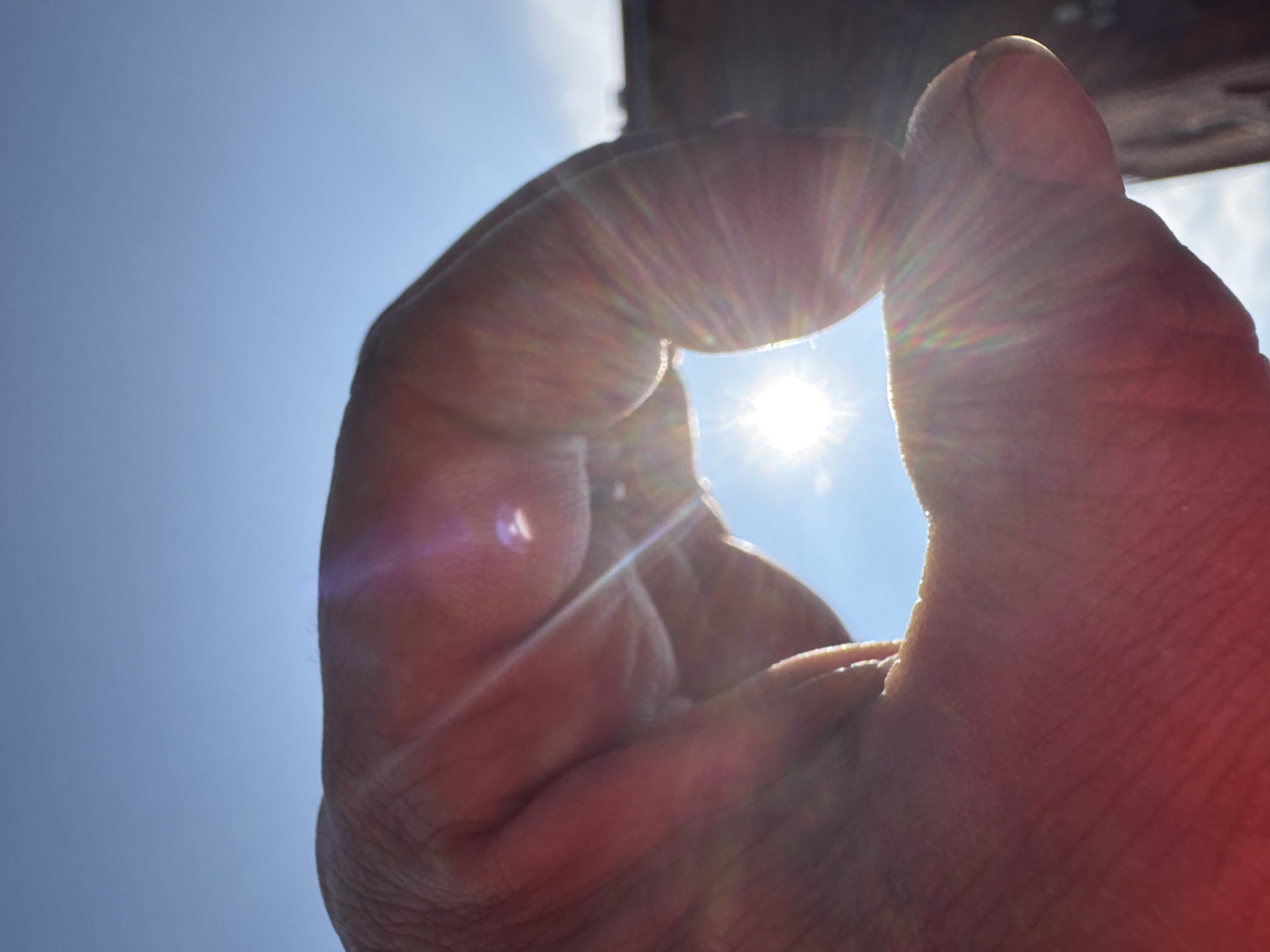 A person's hand is held up against a bright blue sky, with the sun shining through a gap formed by their thumb and index finger. The sun appears as a bright, starburst-like shape with visible lens flares radiating outwards. The hand is close to the camera, and its texture, including wrinkles and skin pores, is visible. The background shows a blurry dark structure, possibly a building, on the right side. The overall impression is a sunny day with a creative framing of the sun. The image was taken in Verkh-Sosnova, Russia.