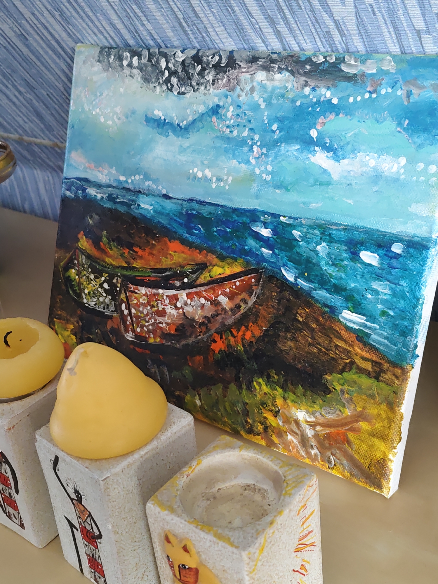 A painting of two boats on a shore is displayed leaning against a blue and white striped wall. The painting depicts a turbulent sea with whitecaps under a sky filled with white and gray clouds and blue patches. The shore is rendered in dark brown and green tones with orange and yellow accents, and two boats, one predominantly red and white and the other green, are visible on the beach. In the foreground, three white, block-shaped candle holders are arranged. The leftmost holder has a yellow candle with a black wick. The middle holder also has a yellow, pear-shaped candle. The rightmost holder is empty. Each candle holder is decorated with an image of a person or animal, rendered in a simple, stylized manner with black lines and red and yellow coloring. The scene appears to be indoors, with a light-colored surface beneath the candle holders and painting. There is no indication of the city of Rostov-on-Don, Russia.