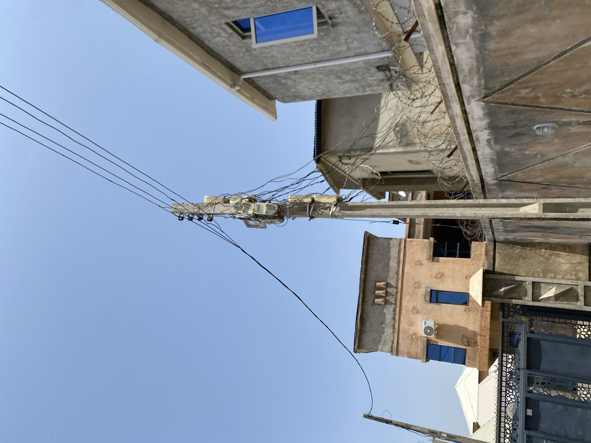 In the city of Jos, Nigeria, this image captures a bright, clear sky over a street scene dominated by residential buildings and utility infrastructure. The primary focus is a concrete utility pole, heavily laden with multiple electrical meters and a complex network of wires, suggesting a densely populated area. Several buildings are visible, including a pale orange structure with blue-tinted windows and an air conditioning unit, and a gray building to its right with a similar blue window. A barbed wire fence tops a concrete wall in the foreground, partially obscuring a dark metal gate with ornate patterns. Another, more distant utility pole is also visible. The overall impression is one of urban development with visible signs of electrical distribution and security measures.
