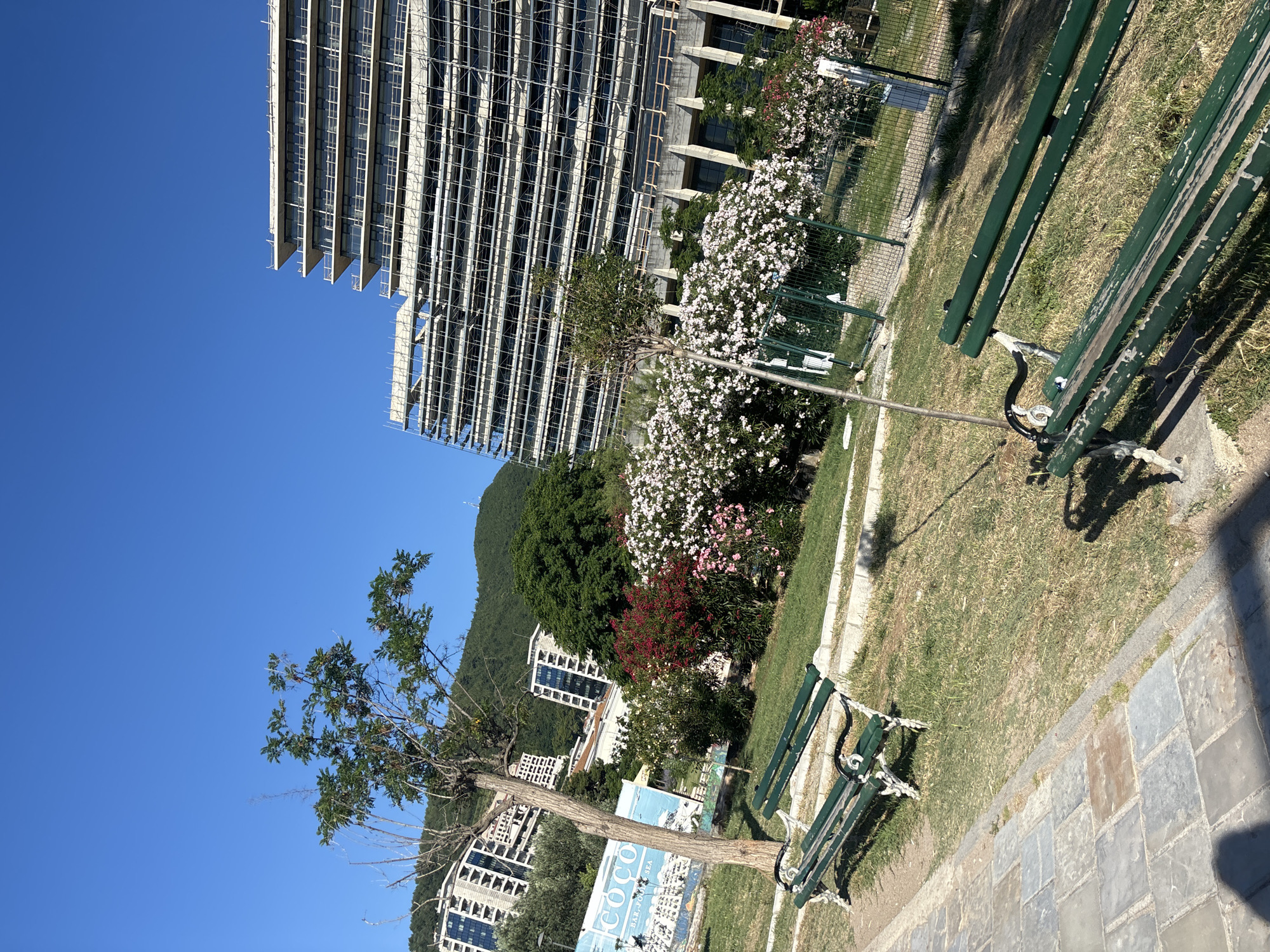The image depicts a sunny day in a park area, likely in Budva, Montenegro, with a large building under construction in the background. The park features a pathway paved with stone tiles, bordered by a strip of grass. Several green park benches are placed on the grass, with one in the foreground showing signs of wear. Lush green grass covers most of the park area, with some patches of dry grass also visible. Blooming bushes with white and red flowers add color to the scene, and a young tree stands near the pathway. A tall building, partially covered in scaffolding, dominates the background, suggesting ongoing construction or renovation. Several other buildings, some appearing residential, are nestled on the hillside behind the park. The sky is a clear, bright blue, indicating fair weather. The overall impression is of a public outdoor space undergoing development, with elements of natural beauty and urban modernization coexisting.