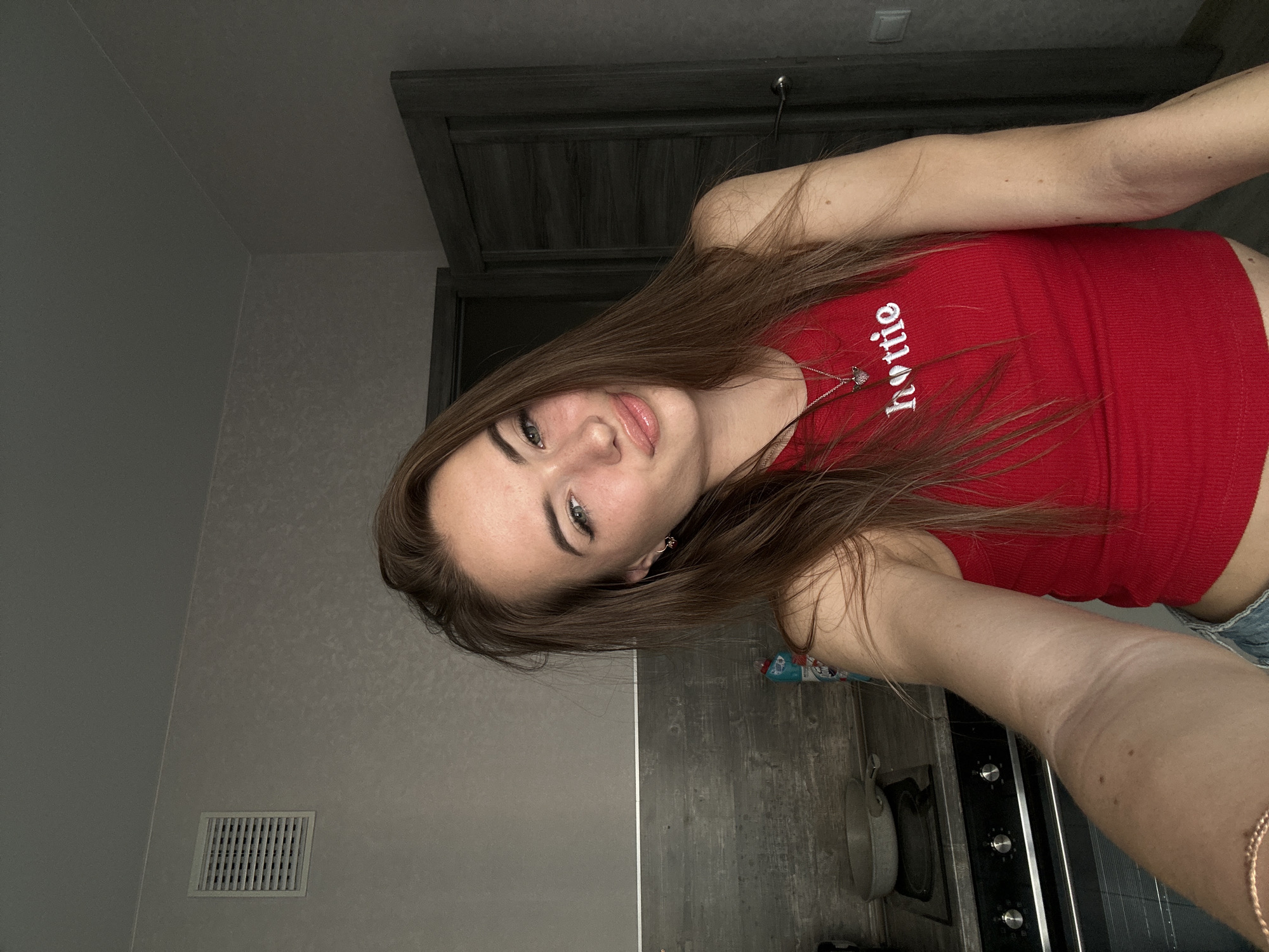 A young woman with long brown hair and blue eyes is taking a selfie in a kitchen. She is wearing a red tank top with the word "hottie" in white letters. The kitchen has dark grey cabinets, a grey backsplash, and a black stovetop with silver knobs. A silver frying pan is on the stovetop. There is a white ventilation grate on the wall to the left of the woman. A dark grey door is visible in the background to the right. The lighting in the room suggests it is daytime.