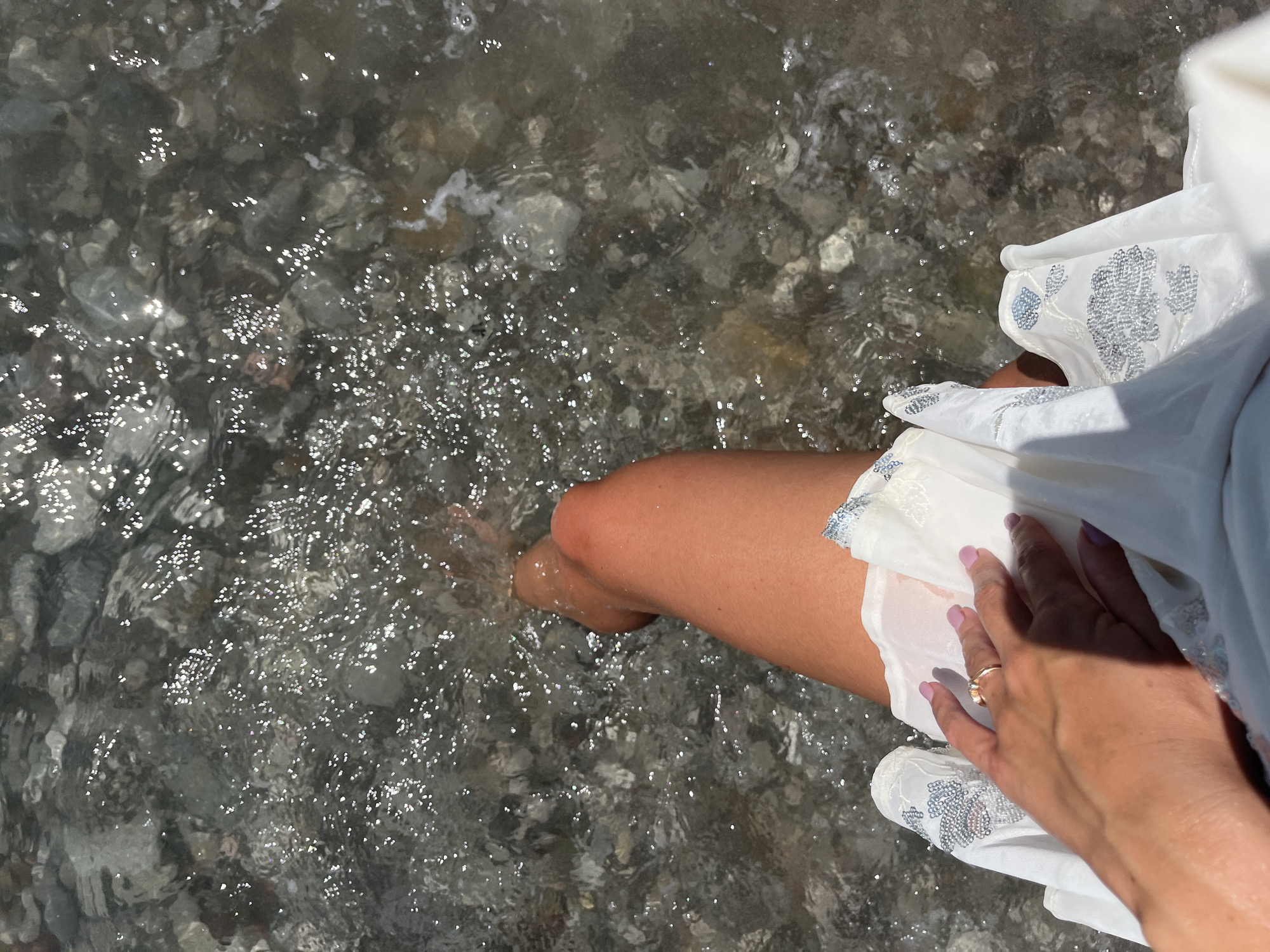 A close-up overhead shot shows a person's legs and hand in the shallow, clear waters of the Adriatic Sea. The person is wearing a white dress adorned with silver embroidery. Sunlight glints off the water's surface, illuminating the rocky seabed beneath. The scene is likely at a beach in Bar, Montenegro.