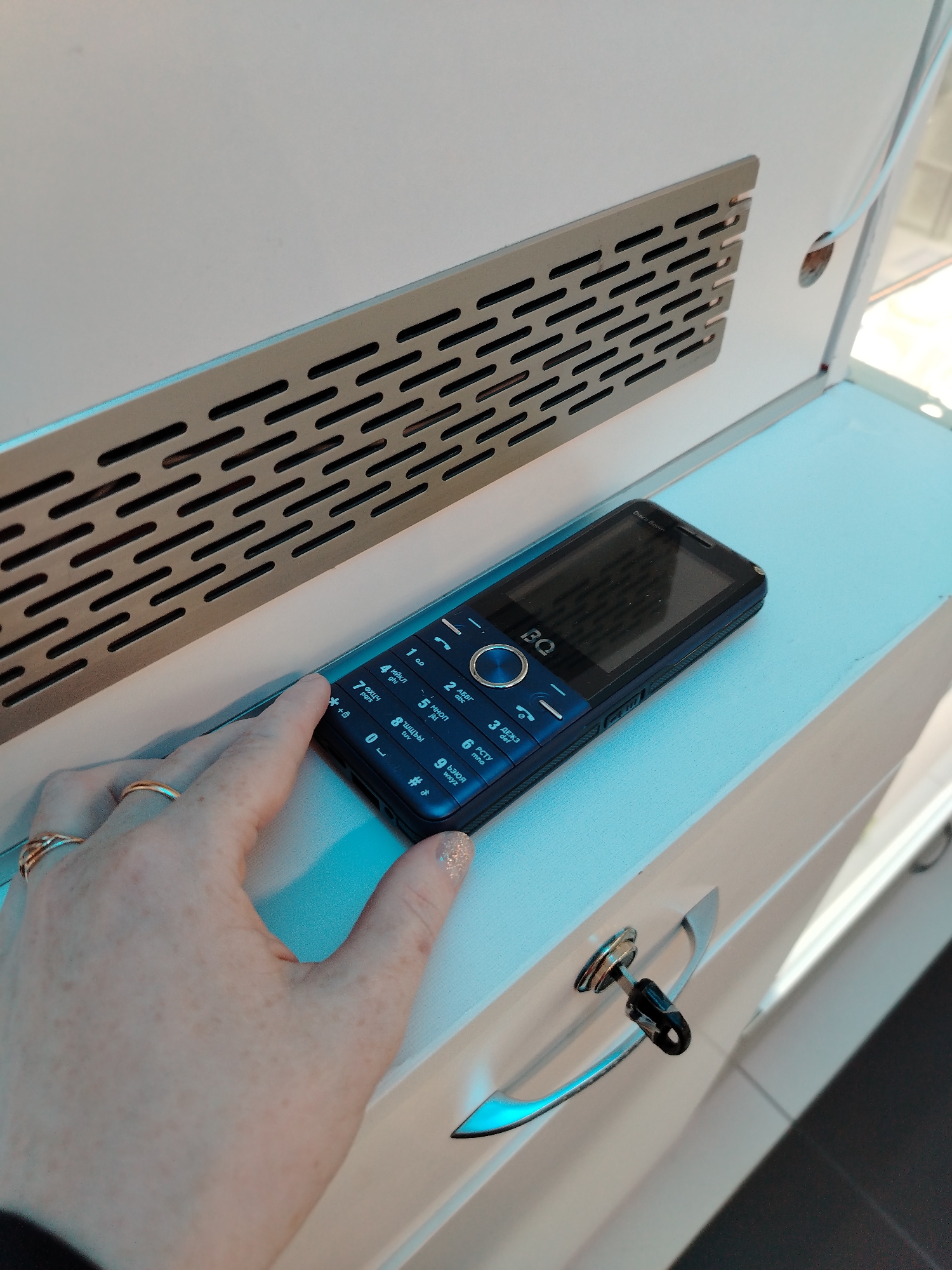 The image shows a black and blue BQ Disco Boom mobile phone placed on a white surface next to a metallic vent. A hand with rings on the fingers is seen touching the phone's keypad. A drawer or cabinet with a silver handle and a key in the lock is visible in the lower right corner. The specific location of Izhevsk, Russia, cannot be determined from the information provided in the image.