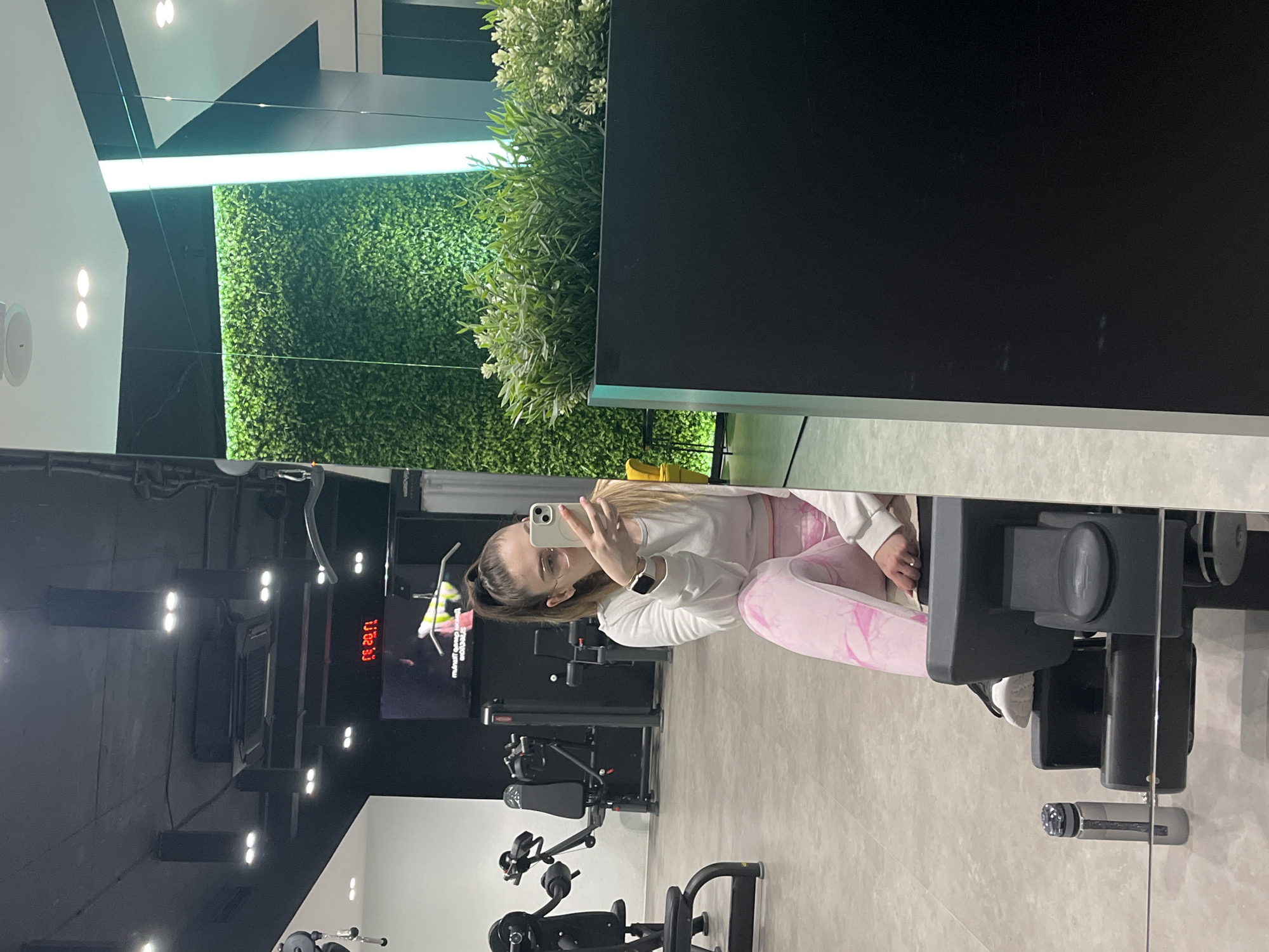 A vertical, eye-level shot shows the reflection of a woman in a gym mirror. The woman has her hair pulled back and is wearing eyeglasses, a white long-sleeve shirt, and pink leggings. She is holding up a smartphone with a white case, apparently taking a selfie. The reflection also captures gym equipment, including weight machines and a screen displaying the time "13:32:33". The backdrop includes a wall covered in green foliage, and a black structure with a plant on top. The location is Unknown, Unknown.