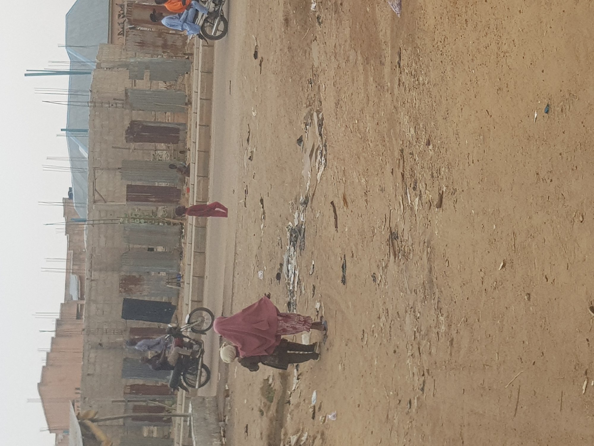The image depicts a street scene in what appears to be Dabawa, Nigeria. The environment is dusty, with unpaved ground dominating the foreground and a paved road to the left. Litter and debris are scattered across the ground. Several partially constructed buildings line the left side of the image. At least three people are visible: one child wearing red, and a person riding a motorcycle with another person. Another person rides a motorcycle in the distance. The overall scene suggests a developing or lower-income urban area.
