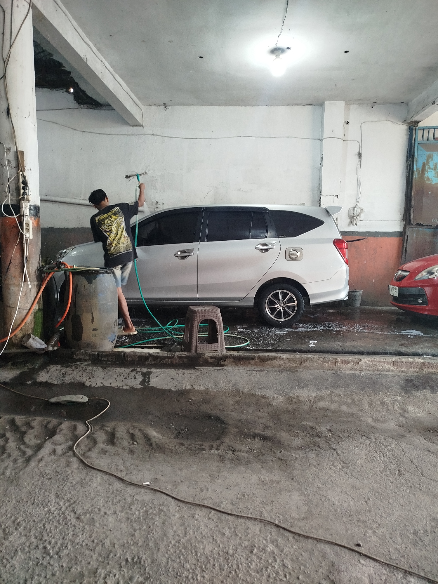 A person is washing a silver car at an outdoor car wash station in Sidoarjo, Indonesia. The car wash appears to be in an industrial or semi-open area, indicated by the concrete floor, exposed ceiling beams, and unpainted walls. The person, wearing a black t-shirt with a graphic design and shorts, is holding a hose and spraying the car from the rear passenger side. A large, gray plastic barrel is positioned next to the car, likely used for water storage. Various hoses and electrical cords are visible on the ground, suggesting the presence of water pumps and other equipment. A small, brown plastic stool is placed near the car. Another red car is parked to the right of the silver car. The environment is somewhat dilapidated, with cracks on the walls and a generally unkempt appearance.