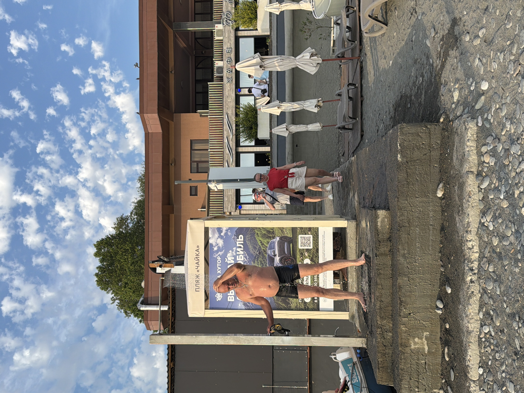 In Sochi, Russia, a man is taking a shower outdoors next to a billboard advertising "ПЛЯЖ «ЧАЙКА»" (Seagull Beach). He is wearing black swim trunks and is holding a dark object, possibly a diving mask, in his left hand. Water is showering down on him from an overhead showerhead. The billboard features an image of a car and text in Russian.
In the background, a building with a brown facade and a sign reading "КАФЕ" (Cafe) is visible. Several closed beach umbrellas and lounge chairs are arranged on the sand. A few people are walking in the background, including a woman in a red top and white shorts and a man in a white t-shirt and shorts. The environment is a beach with a paved walkway and a pebbled shore. The sky is blue with scattered white clouds.