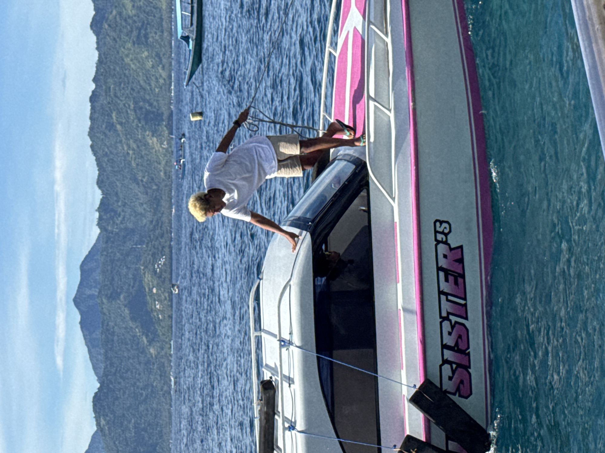 A man stands on a motorboat with "SISTER 'S" written on its side, possibly near Unknown, Indonesia, as a lush green mountain range is visible in the distance. The water is deep blue, with other boats anchored nearby. The man has light-colored hair, is wearing a white t-shirt and tan shorts, and is holding a rope.