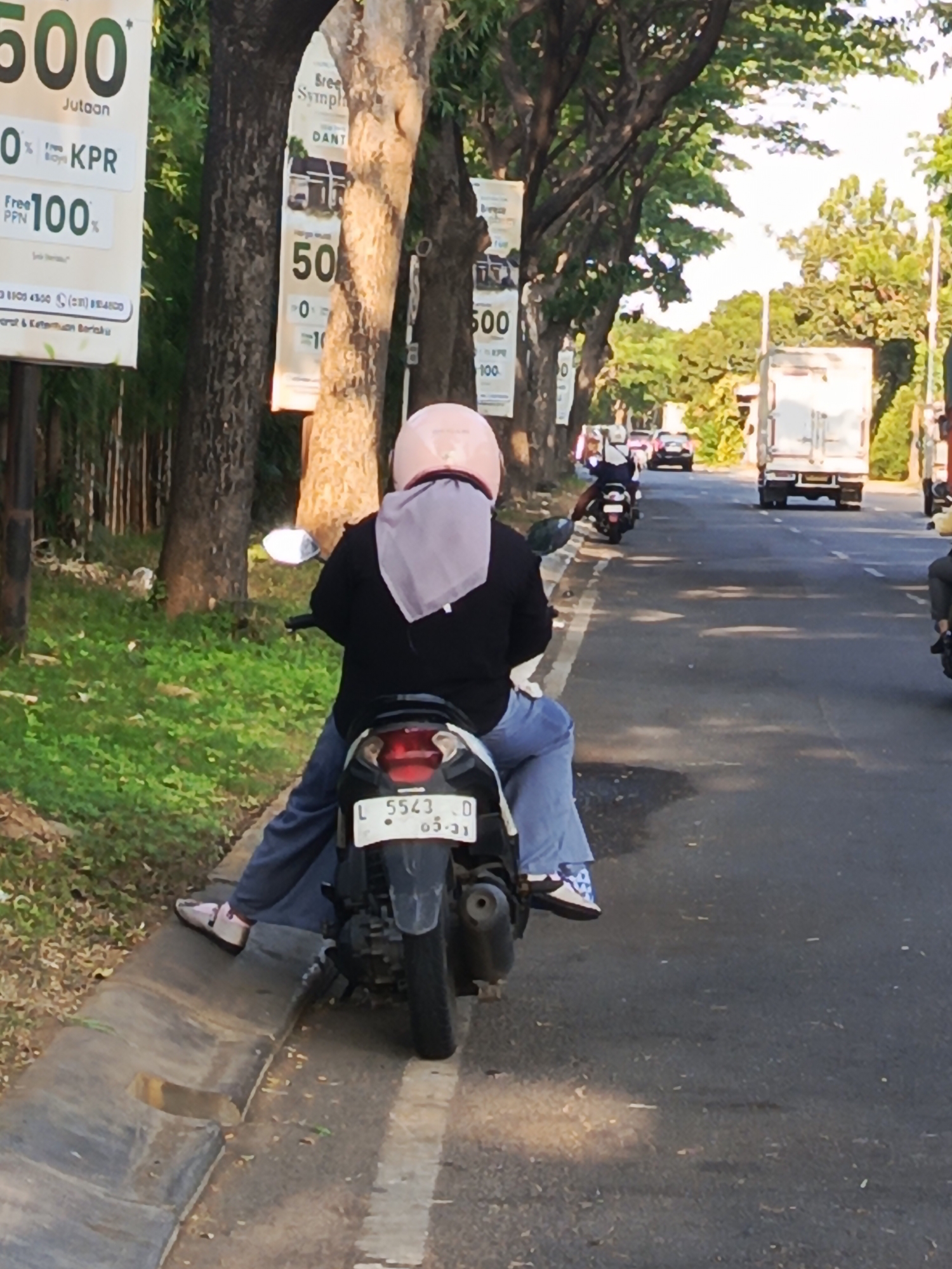 In Calukan RT 1 RW 5 Keboansikep, Indonesia, a person wearing a pink helmet and a light purple headscarf is riding on the back of a motorcycle. They are wearing a dark long-sleeved shirt and loose blue pants. Their feet are resting on the motorcycle's footpegs, and they are wearing white and blue patterned shoes. The motorcycle has a license plate that reads "L 5543 D 05-21". The motorcycle is positioned on the right side of a paved road, with a grassy area and a concrete curb to its left. In the background, there are trees lining the road, some with banners hanging from them advertising "500 Jutaan", "Free KPR", and "Free PPN 100%". Further down the road, other vehicles, including a white truck and other motorcycles, are visible. The scene appears to be during daylight hours, with sunlight casting shadows on the road.