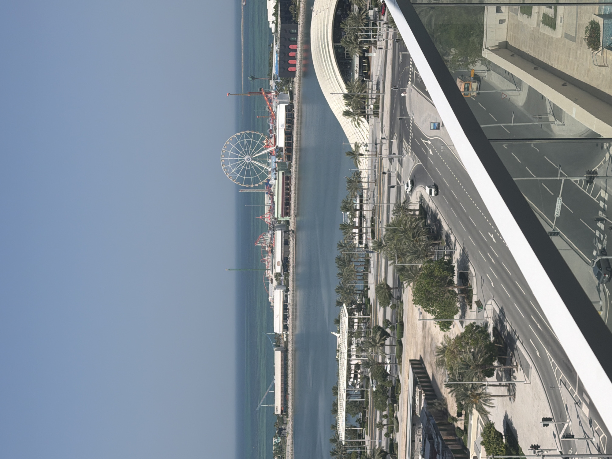 An eye-level, outdoor, vertical, long shot captures a waterfront city district on a clear, sunny day under a pale blue sky. In the background, the turquoise ocean meets the horizon. Across the calm water, a strip of land hosts an amusement park with a large white Ferris wheel, various roller coasters, and buildings with red and black facades. A white, wave-like structure with dark windows dominates the middle ground, situated beside a body of water. In the foreground, a multi-lane road curves around a body of water, bordered by palm trees and other greenery. Several cars and a white van are visible on the road. A balcony railing made of glass and metal is in the lower portion of the image, obscuring a portion of the scene.